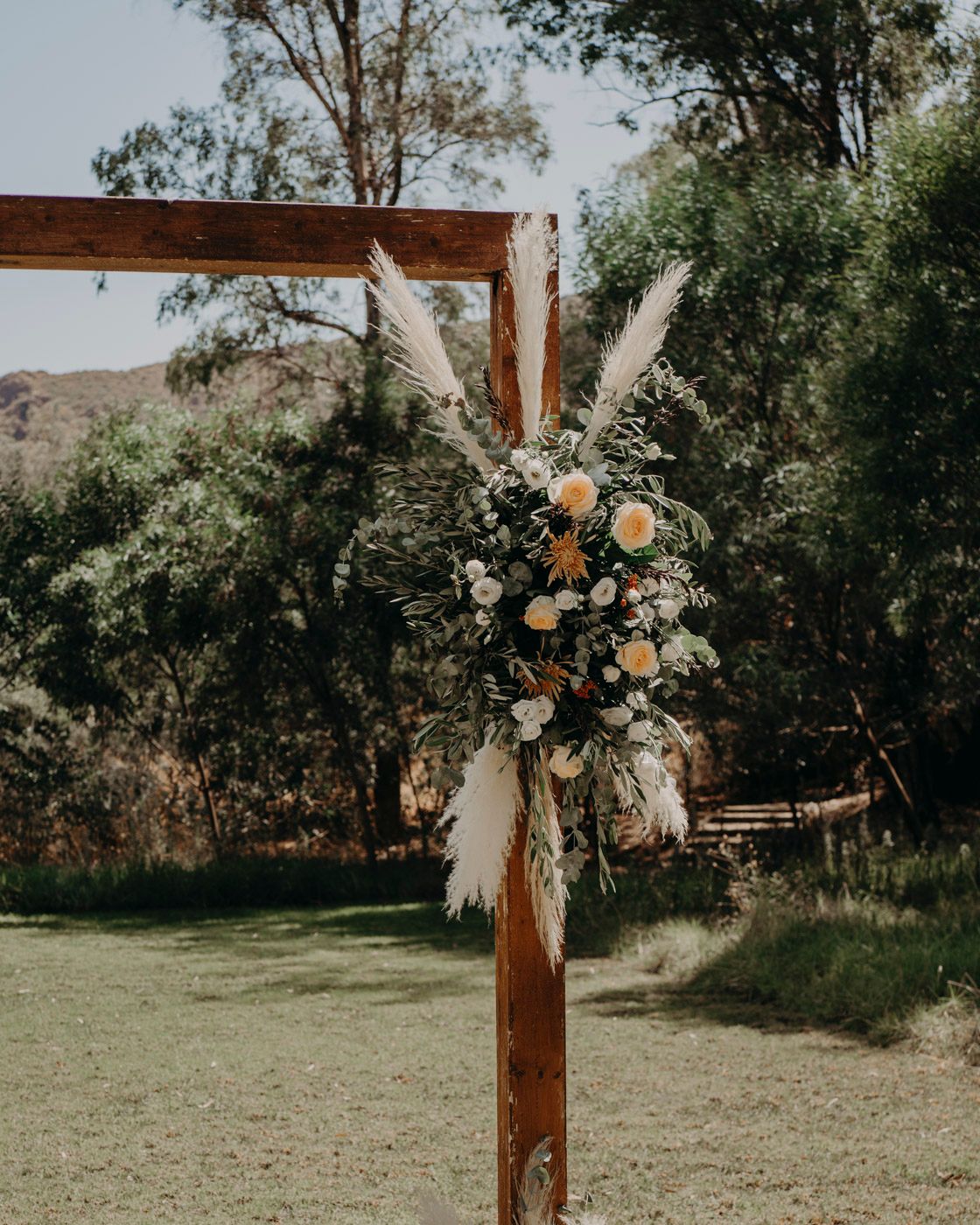 Rustic wooden cross decorated with pampas grass and floral arrangements against a natural outdoor backdrop.