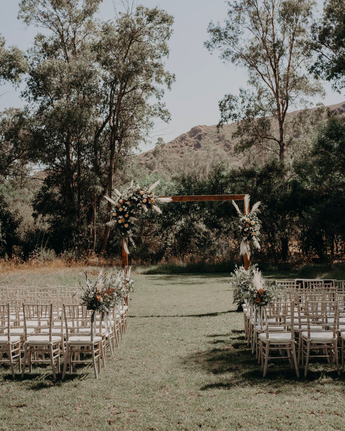 Outdoor wedding ceremony setup with rows of wooden chairs and floral arches leading to a wooden altar in a mountain setting.