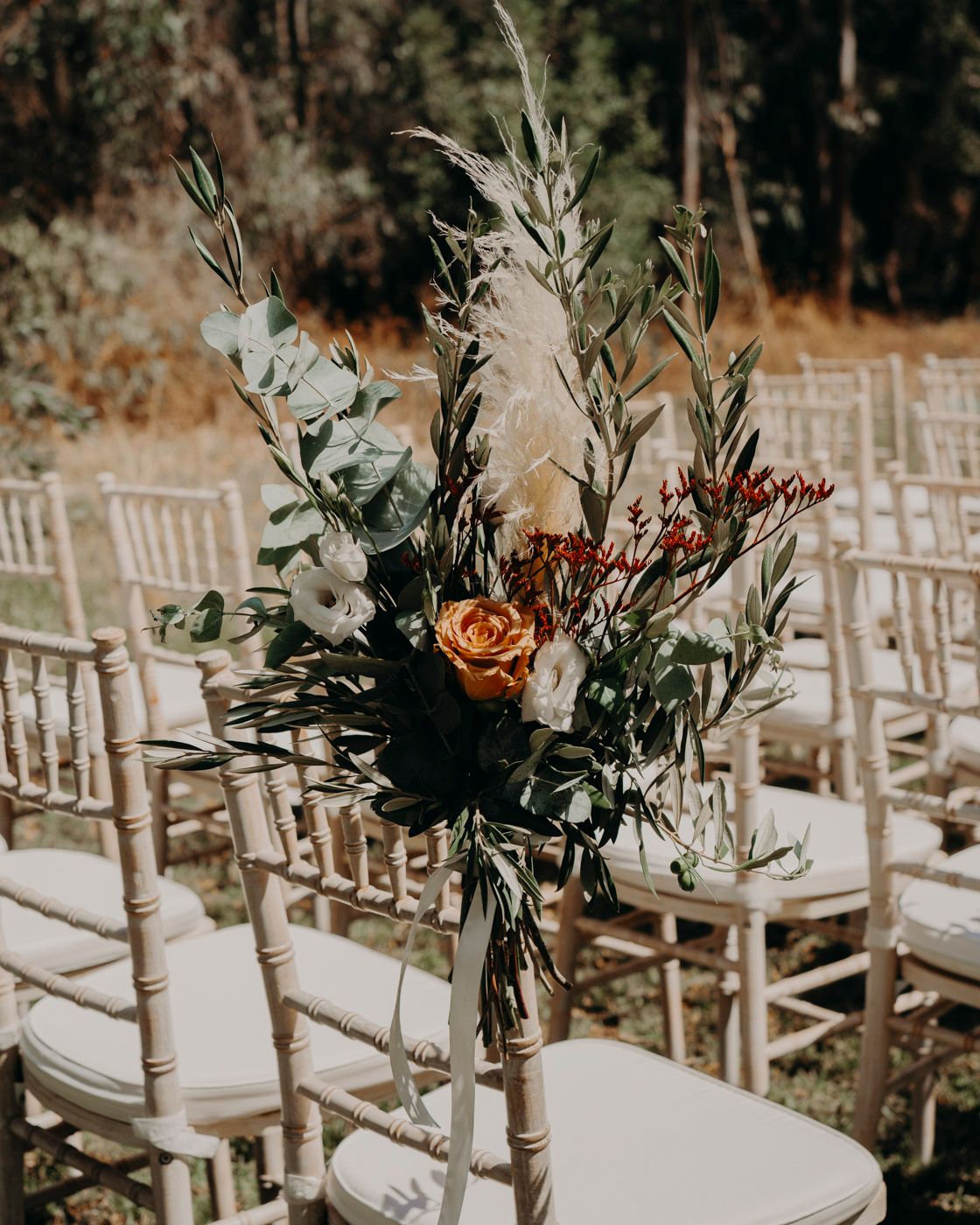 Rustic wedding ceremony setup with white chiavari chairs and elegant floral arrangements in muted tones and pampas grass.