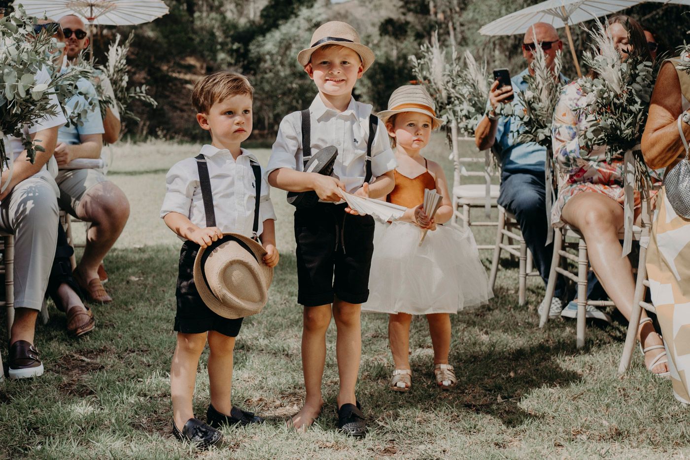Three young children in vintage-inspired attire with suspenders and straw hats at an outdoor wedding ceremony.