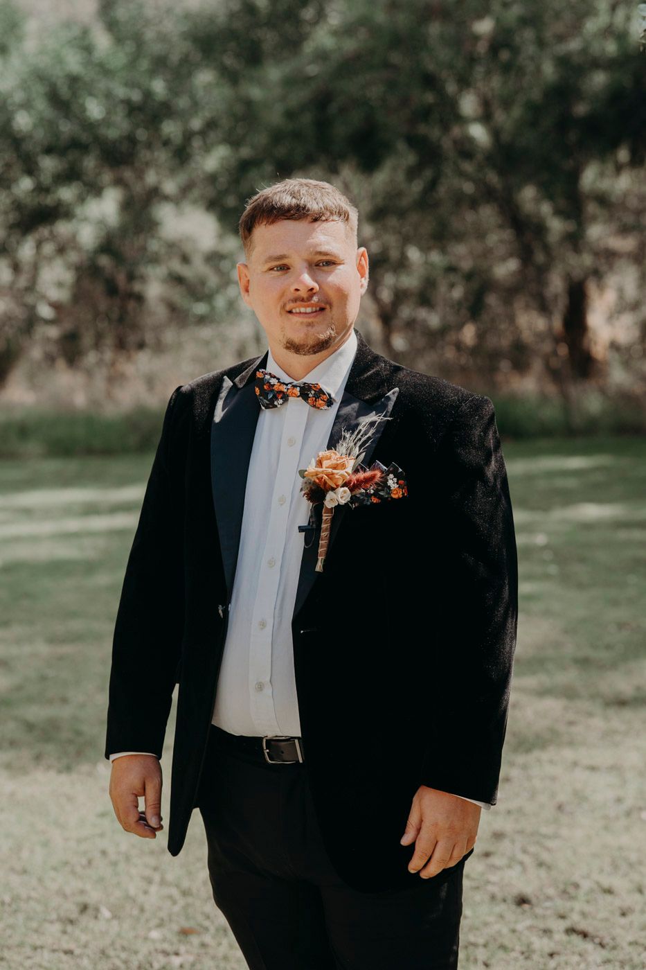 Wedding boutonniere with orange and blush flowers adorns a classic black tuxedo and bow tie in an outdoor setting.