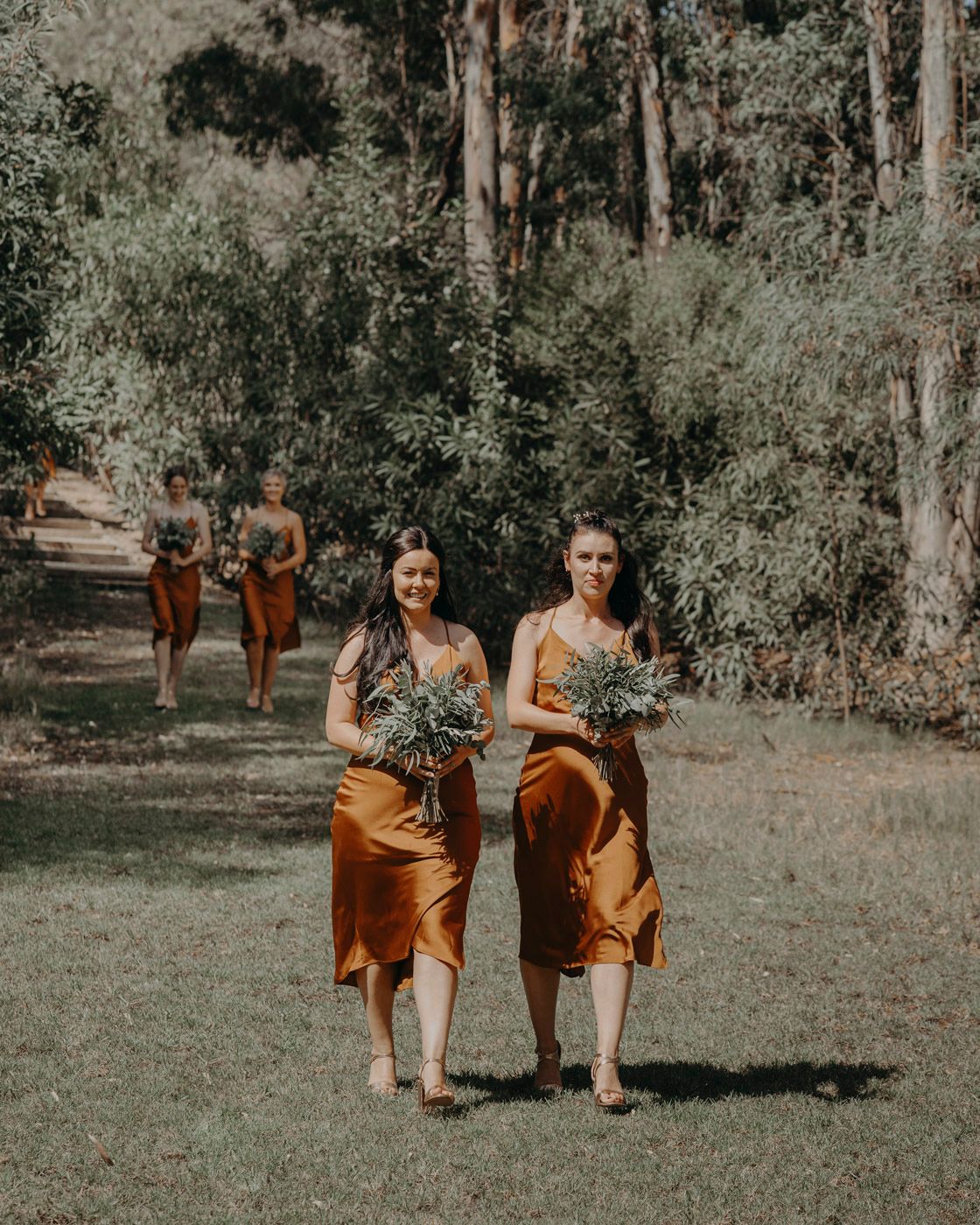 Bridesmaids in rust-colored satin dresses walk together holding bouquets on a wooded path during an outdoor wedding.