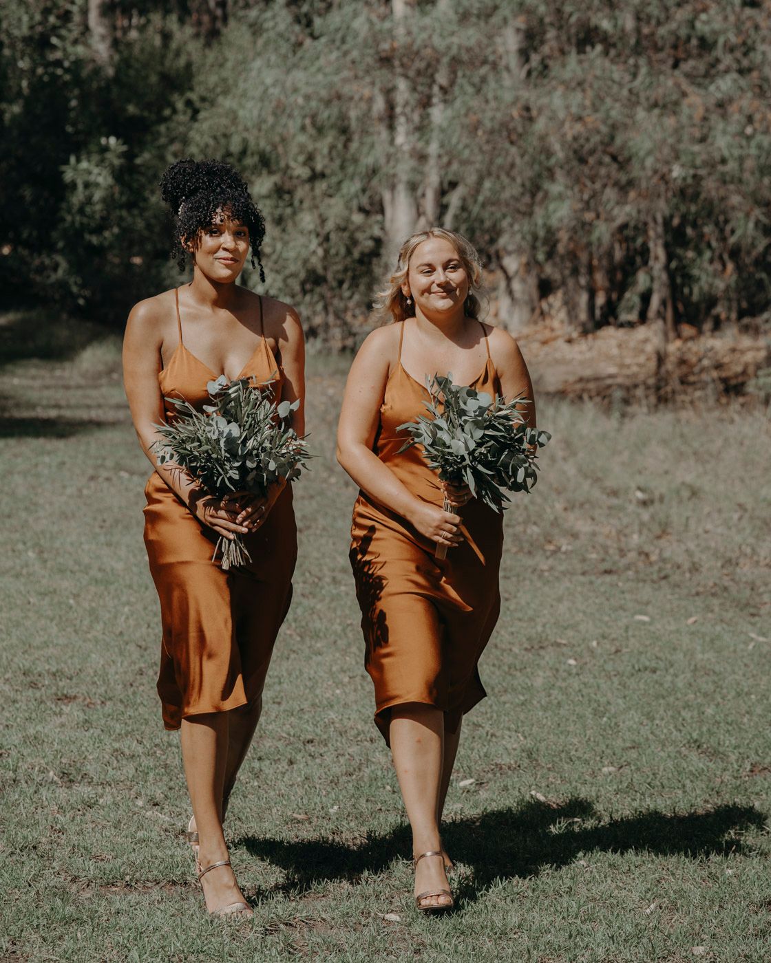 Two bridesmaids in matching copper-colored silk dresses carry greenery bouquets while walking outdoors on a sunny day.