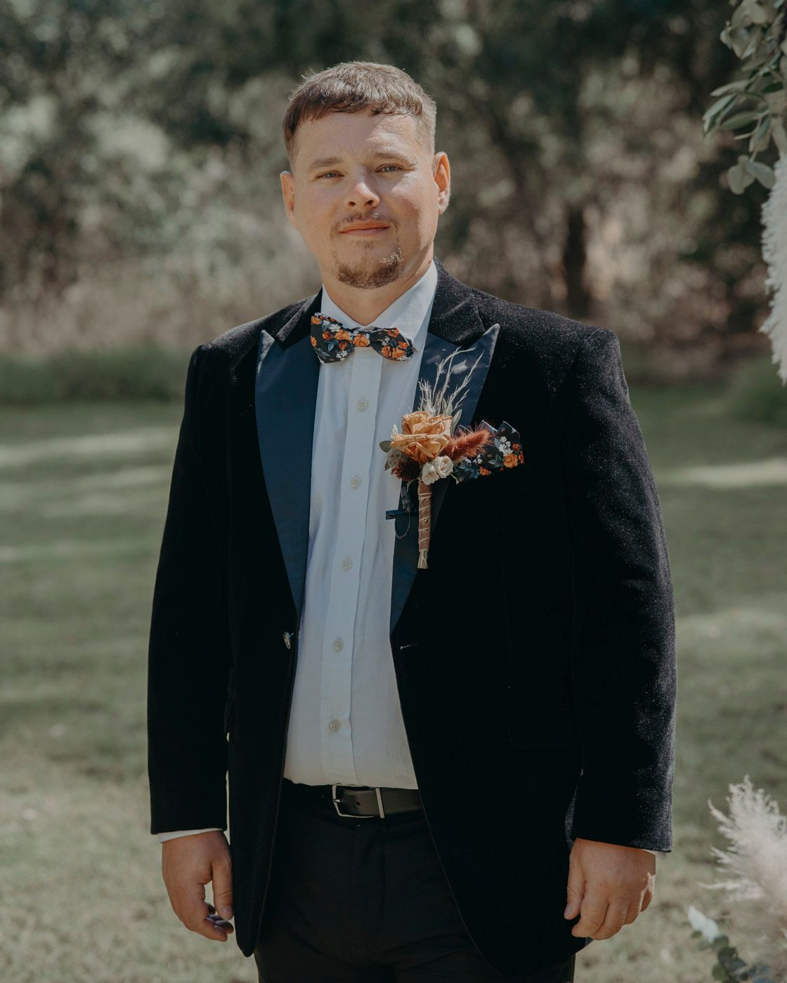 Wedding attendee in black suit and bowtie with orange floral boutonniere poses outdoors in natural setting.