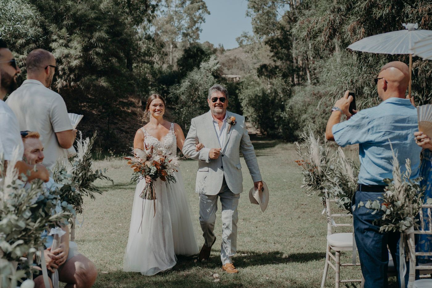 Couple walking down outdoor aisle decorated with greenery during sunny garden wedding ceremony.