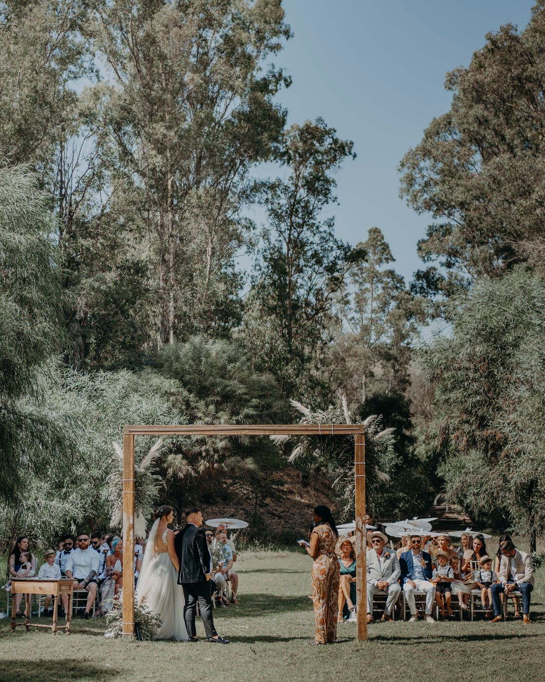An outdoor wedding ceremony under a rustic wooden arch surrounded by tall trees and natural scenery with guests seated.