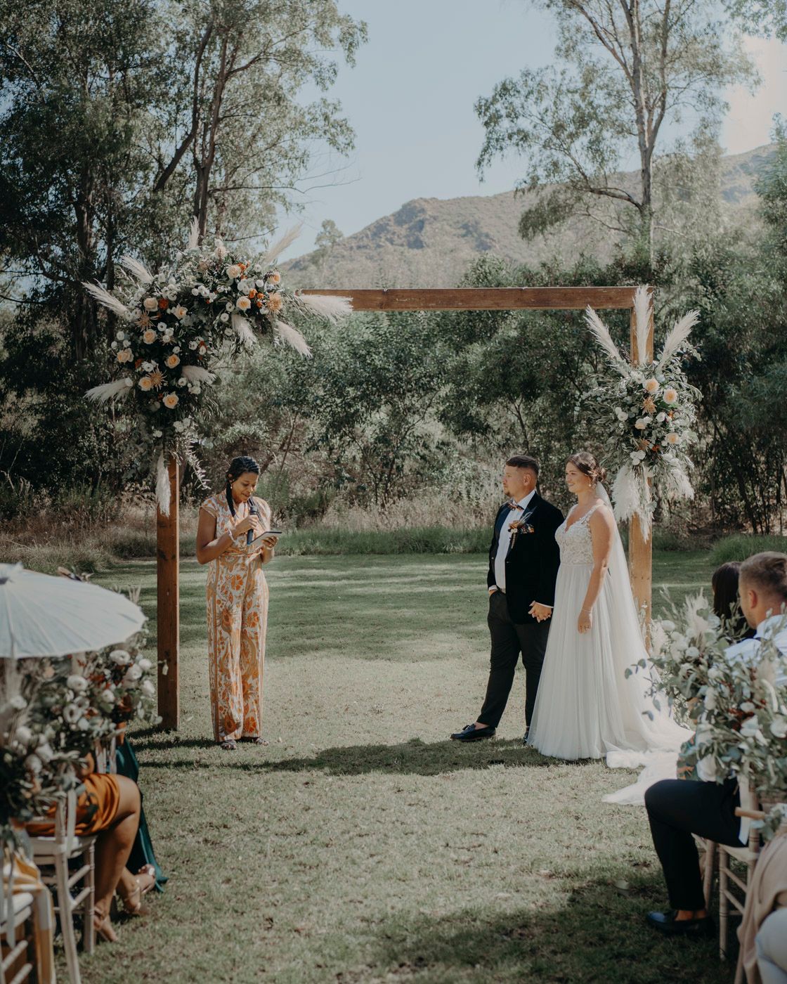 A rustic outdoor wedding ceremony under a wooden arch decorated with floral arrangements in a mountain landscape.