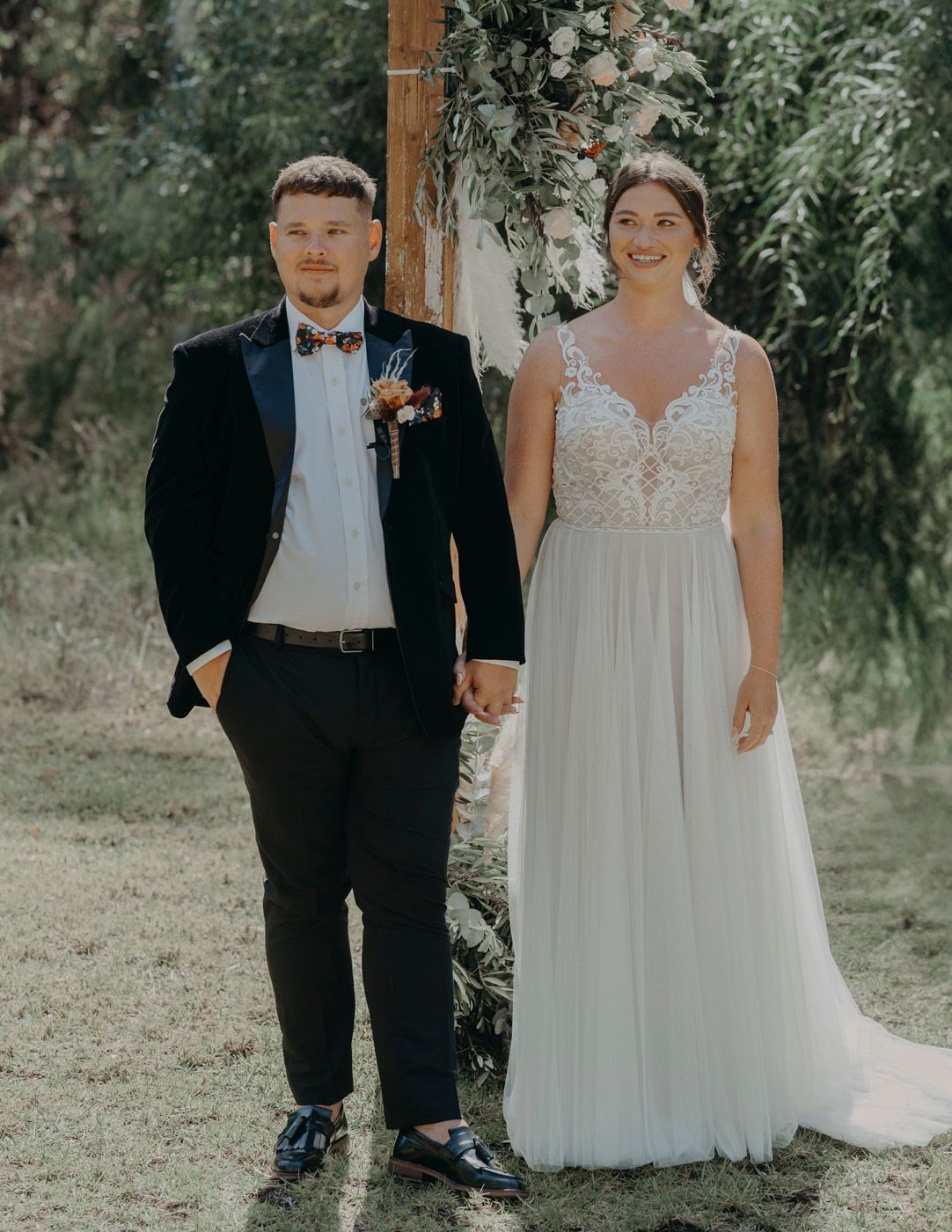 A couple poses outdoors on their wedding day, with the bride in a flowing white gown and groom in classic black tuxedo.