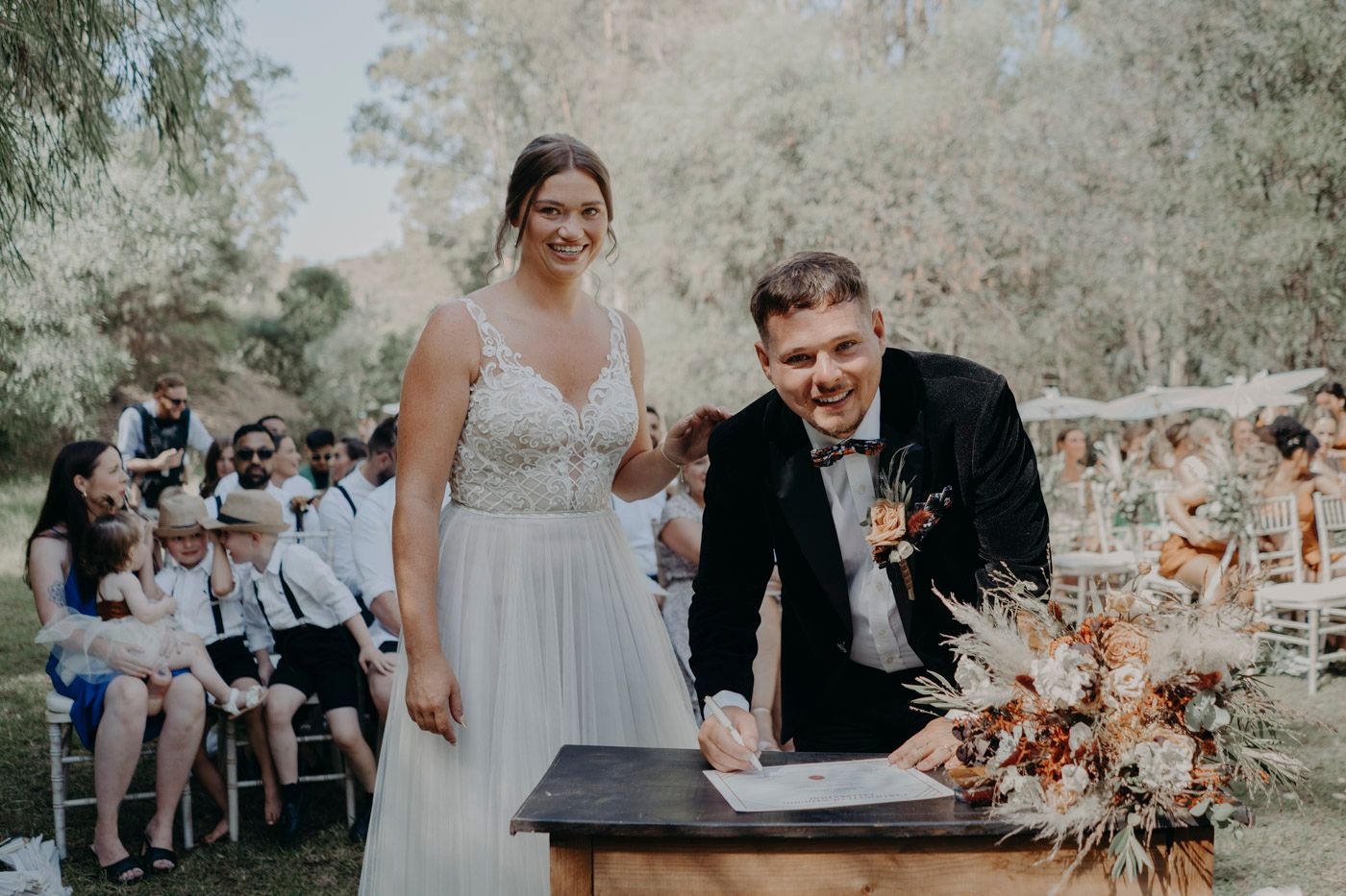 Newlyweds sign marriage documents at outdoor ceremony with guests seated in background and floral decor on table.