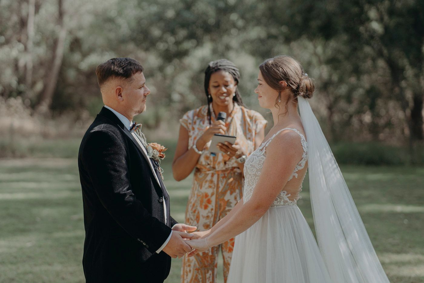 A wedding ceremony takes place outdoors on a sunny day with an officiant and couple holding hands.