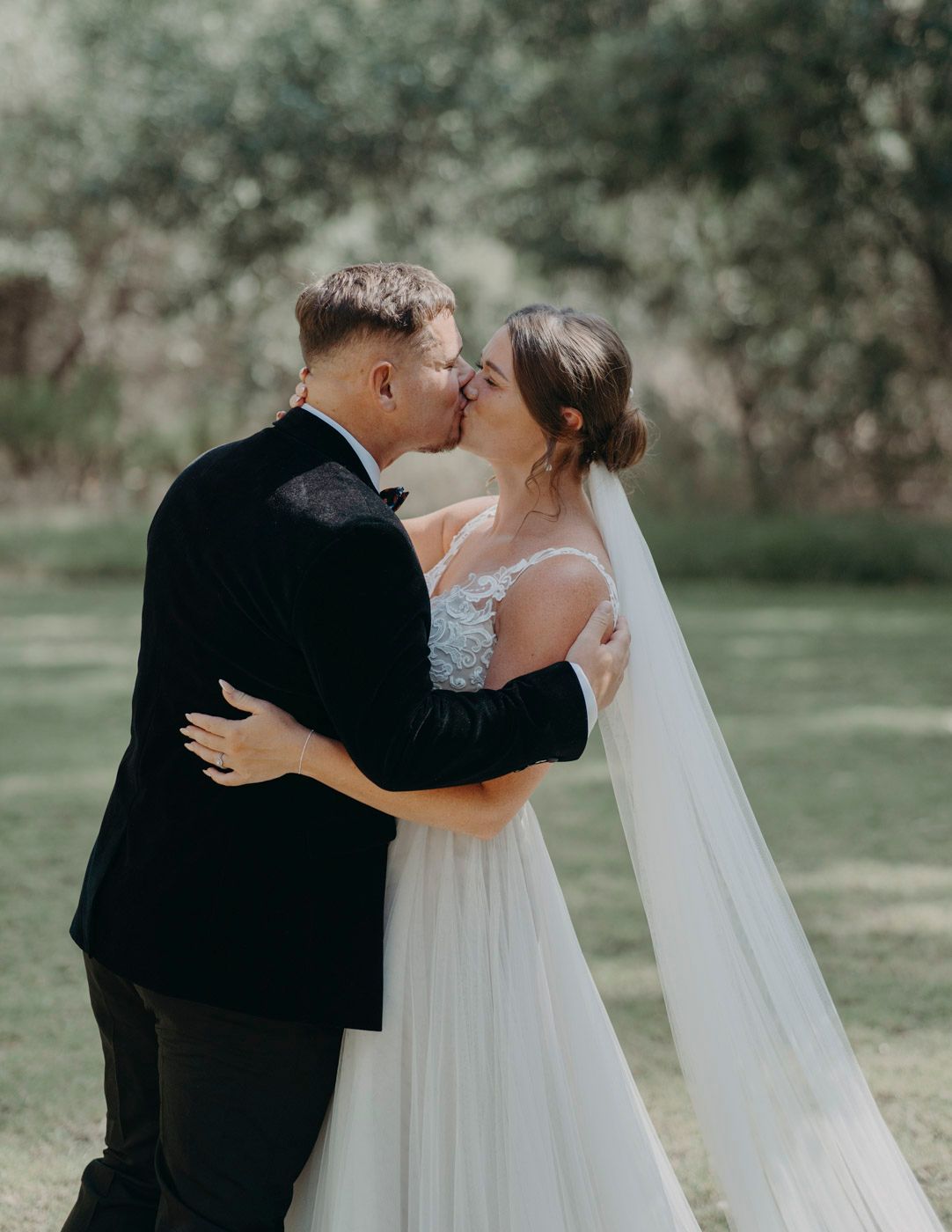 Romantic wedding couple sharing an intimate kiss outdoors with flowing white veil and black suit against natural greenery.