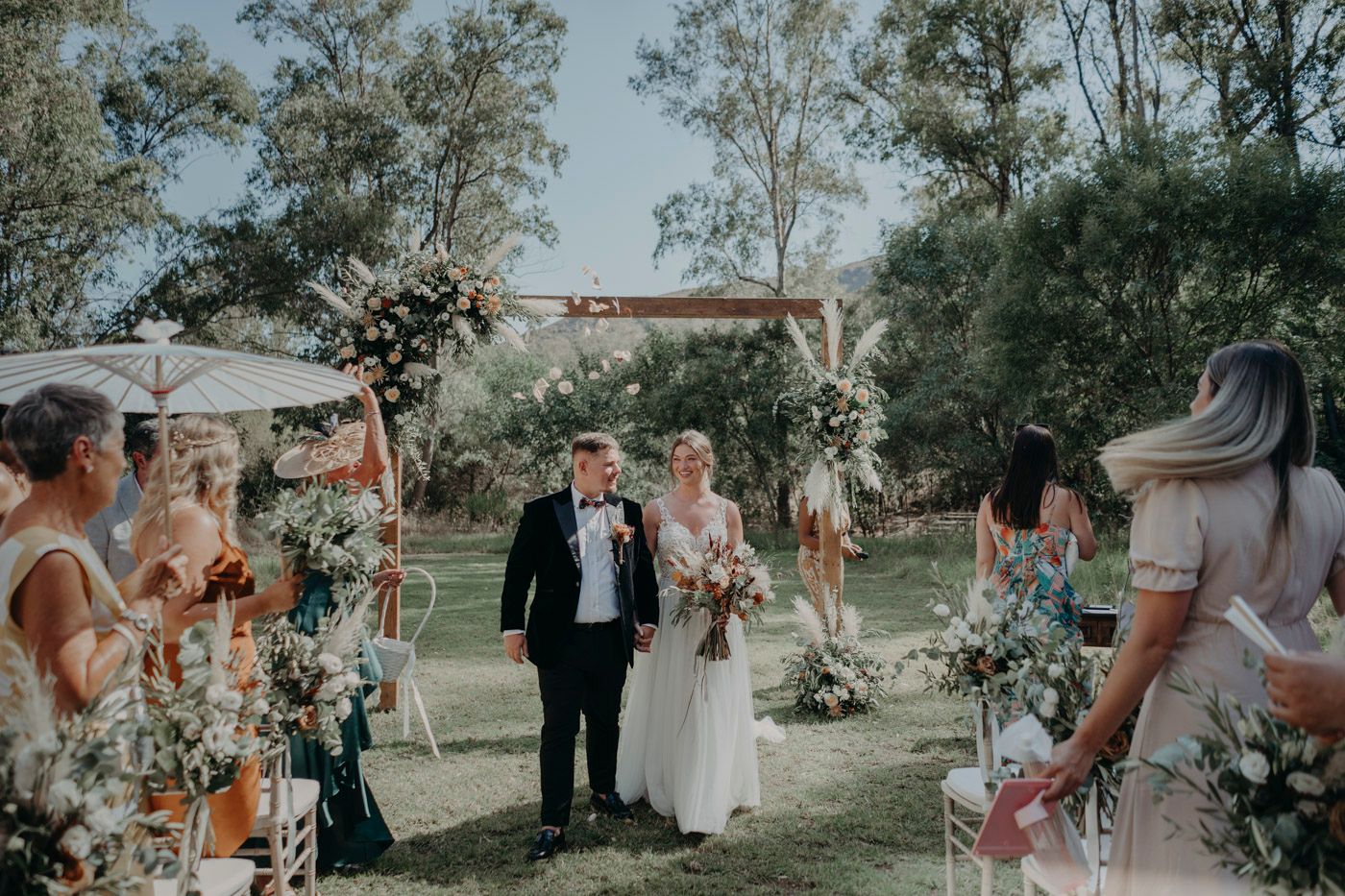 An outdoor garden wedding ceremony takes place under a wooden arch adorned with delicate floral arrangements.
