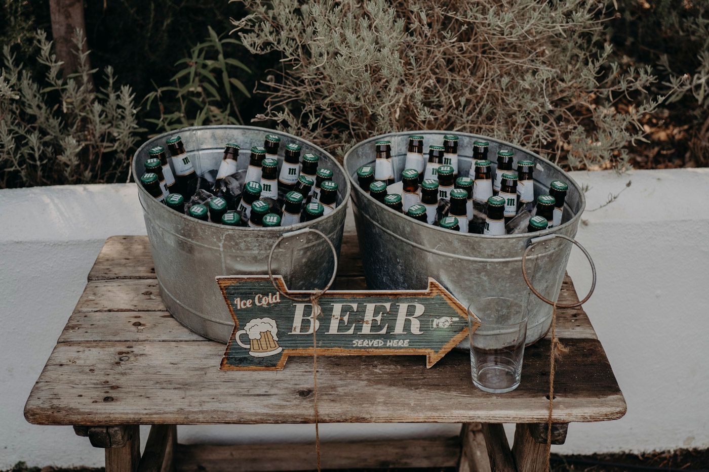 Two metal buckets filled with beer bottles sit on a rustic wooden table with a vintage beer sign and empty glass.