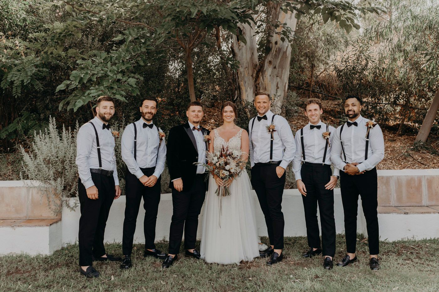 A wedding party in a garden setting with people wearing black pants, white shirts, suspenders, and a white gown.