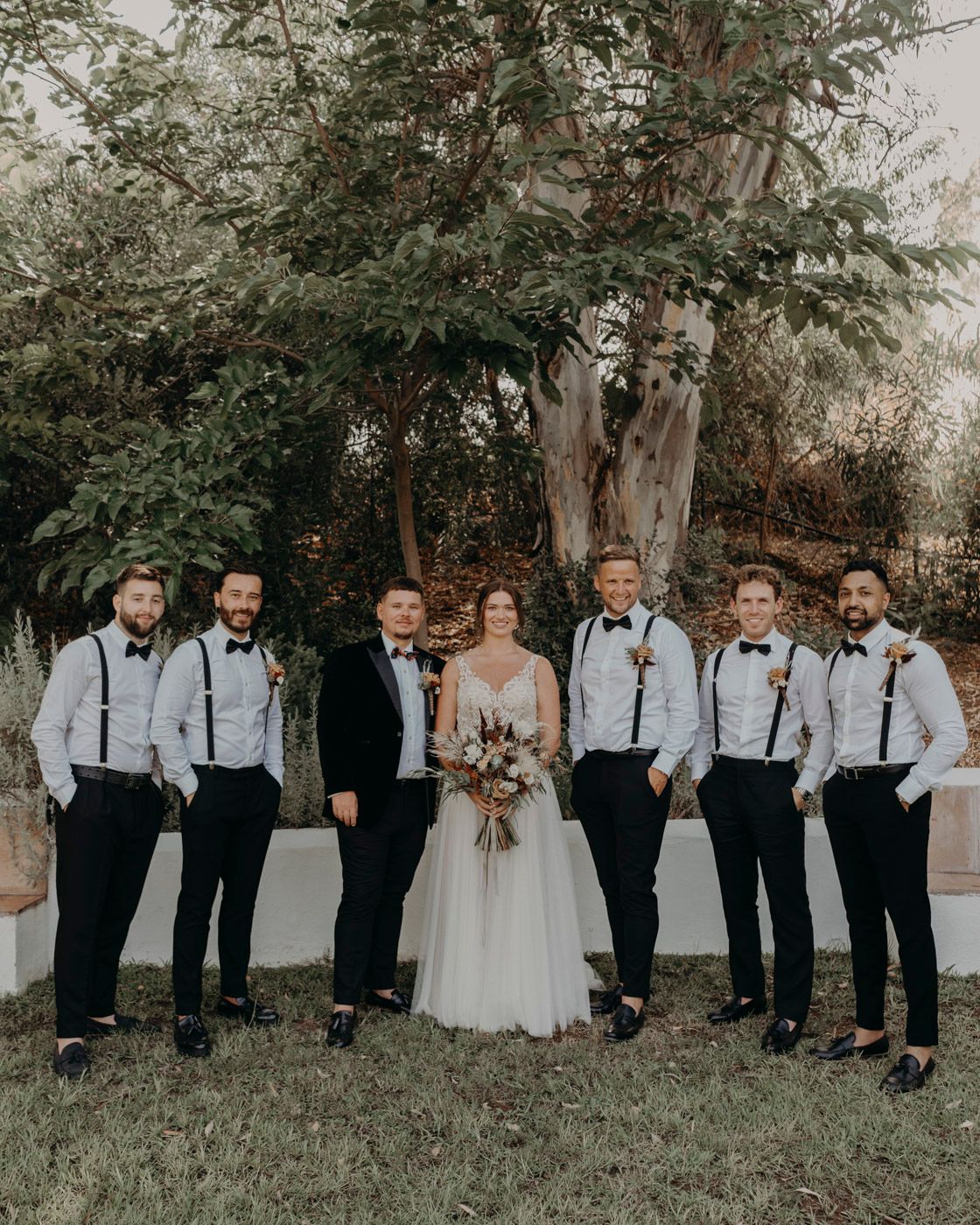 A wedding party in black and white attire poses together outdoors under a leafy tree with a rustic autumn backdrop.