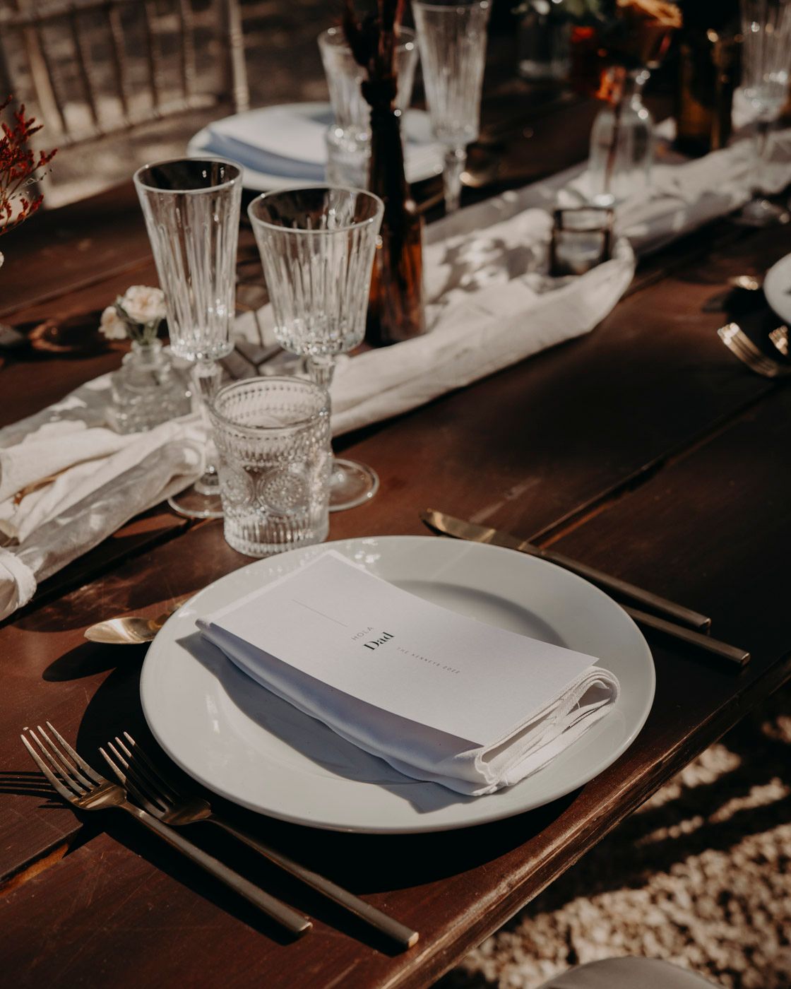 Elegant table setting with crystal glasses, white plates, and table linens on a rustic wooden table in moody lighting.