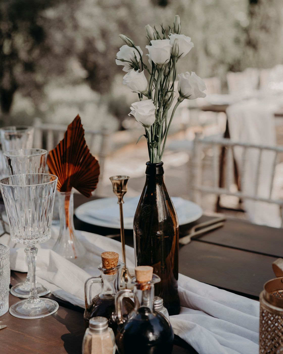 Wedding table decoration with dark bottle vase containing white flowers, crystal glasses and small glass bottles on a wooden table.