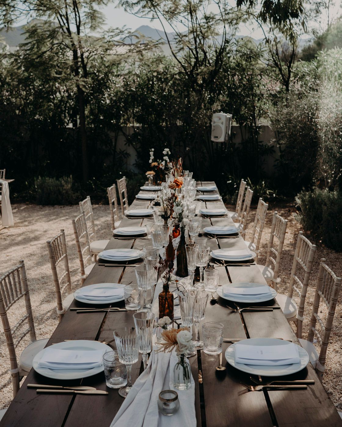 Elegantly set outdoor dining table with rustic chairs and place settings under trees for an intimate garden reception.