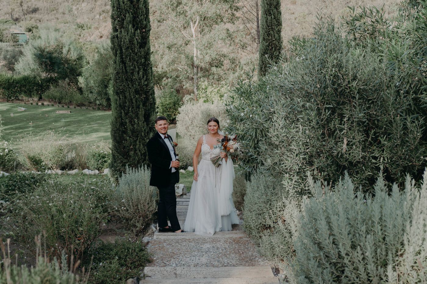 A couple stands together on a garden path surrounded by cypress trees and lush greenery in a rustic outdoor setting.