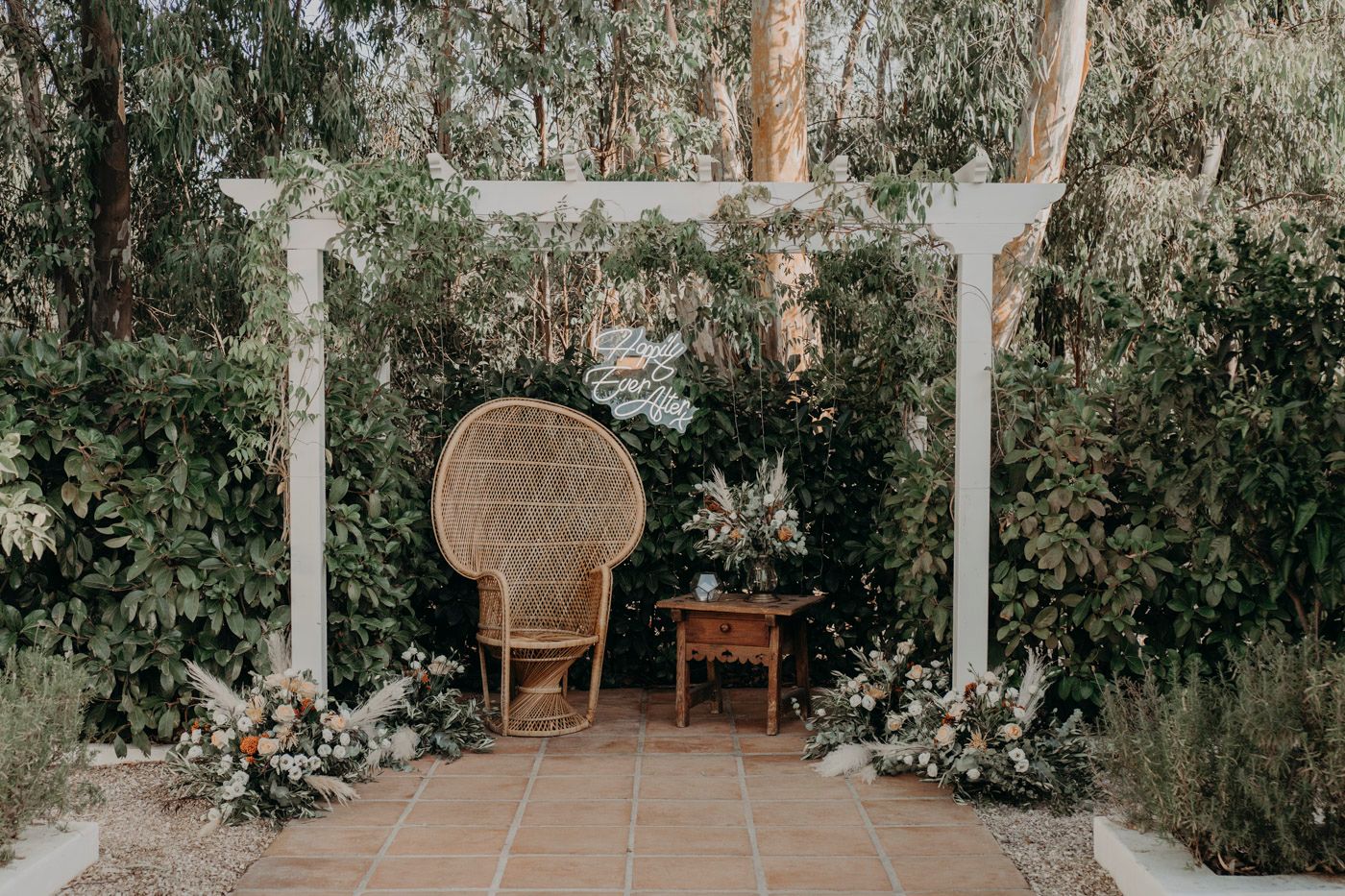 A rustic garden arbor with a peacock chair and small side table adorned with white floral arrangements and greenery.