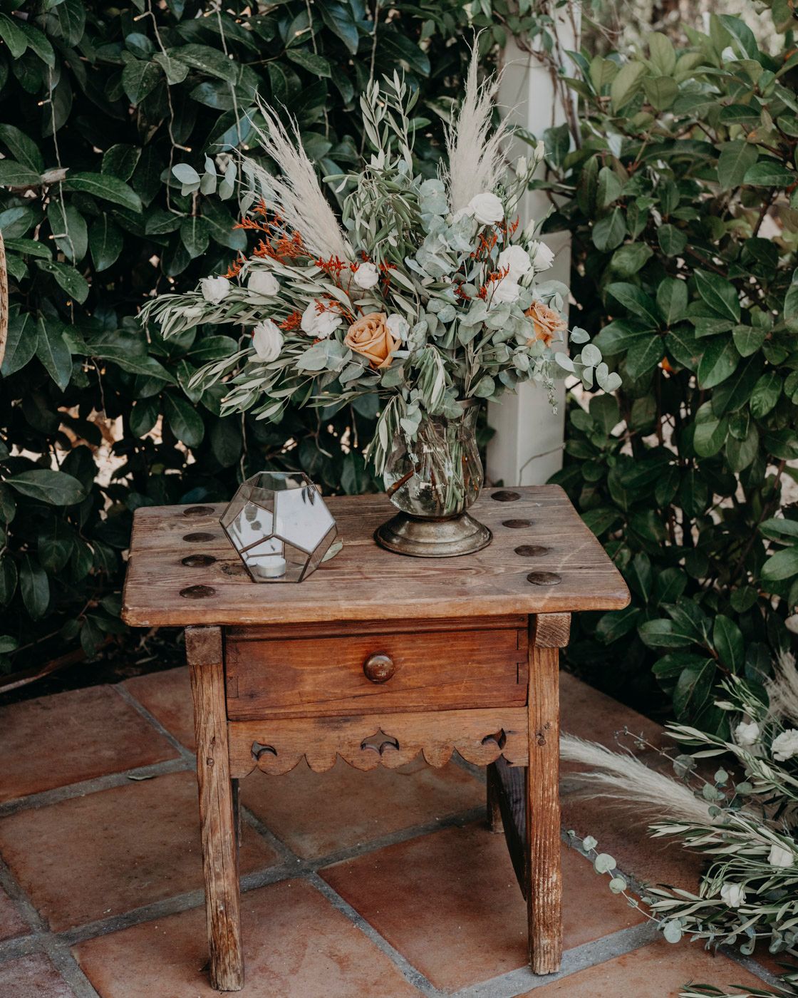 Rustic wooden side table with terracotta tiles displays a geometric candle holder and floral arrangement with greenery.