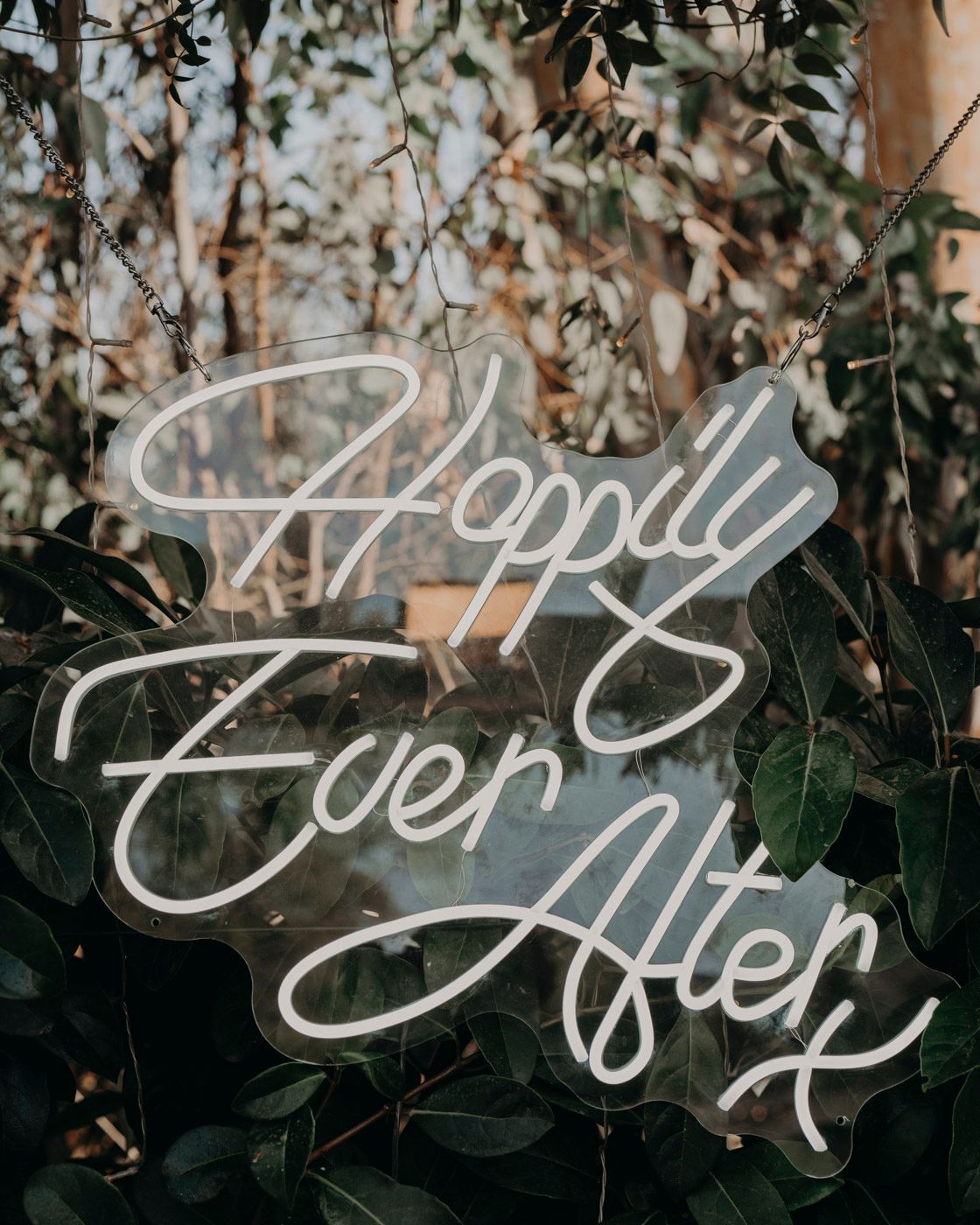 White neon sign reading 'Happily Ever After' mounted against green foliage and leaves in a romantic garden setting.