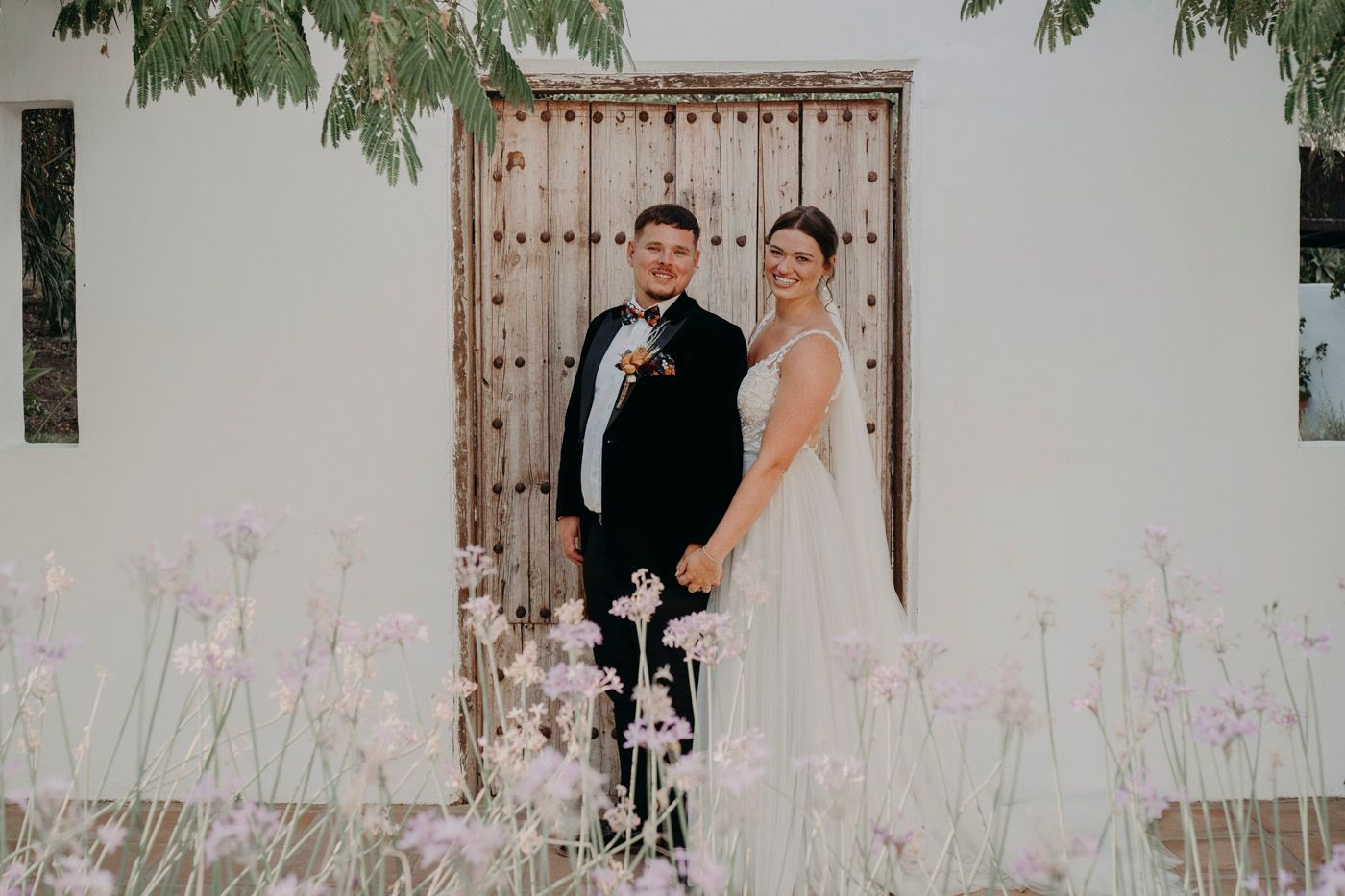 A couple in wedding attire stands in front of a rustic wooden door with white walls and lavender flowers.