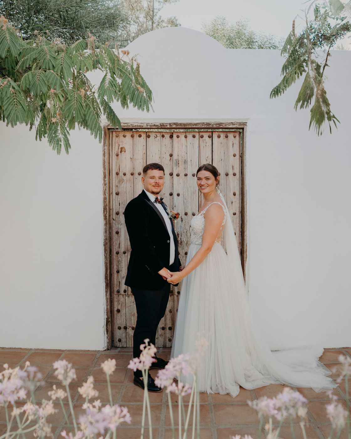 A couple poses against a rustic wooden door on their wedding day, with delicate pink flowers in the foreground.