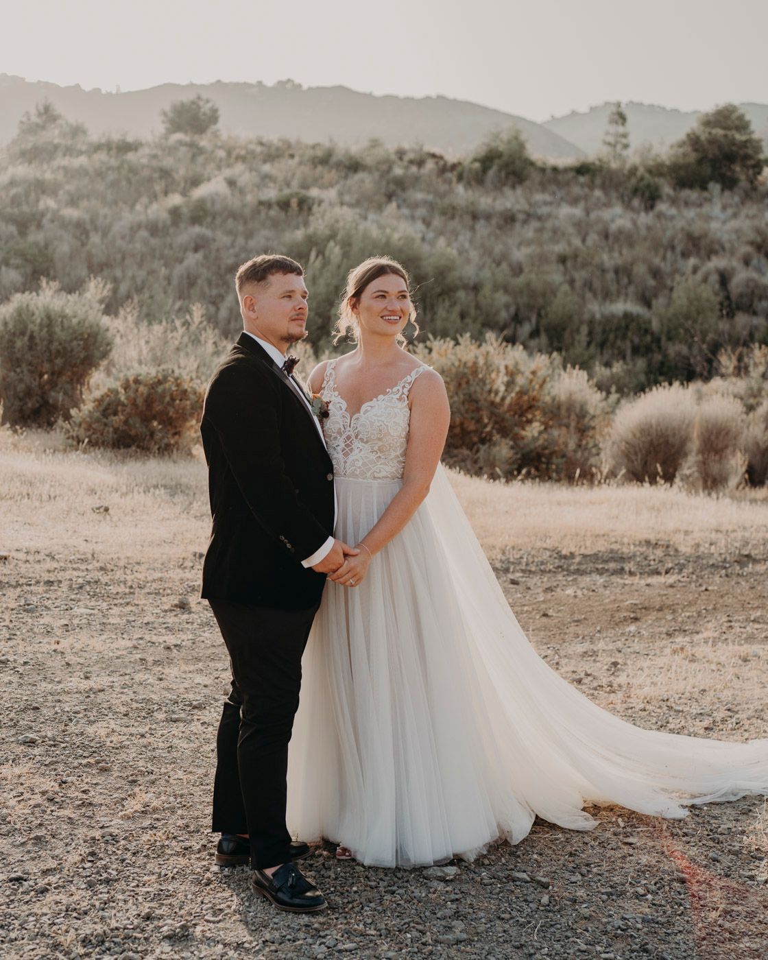 A couple poses together in formal wedding attire against a scenic desert landscape at sunset.