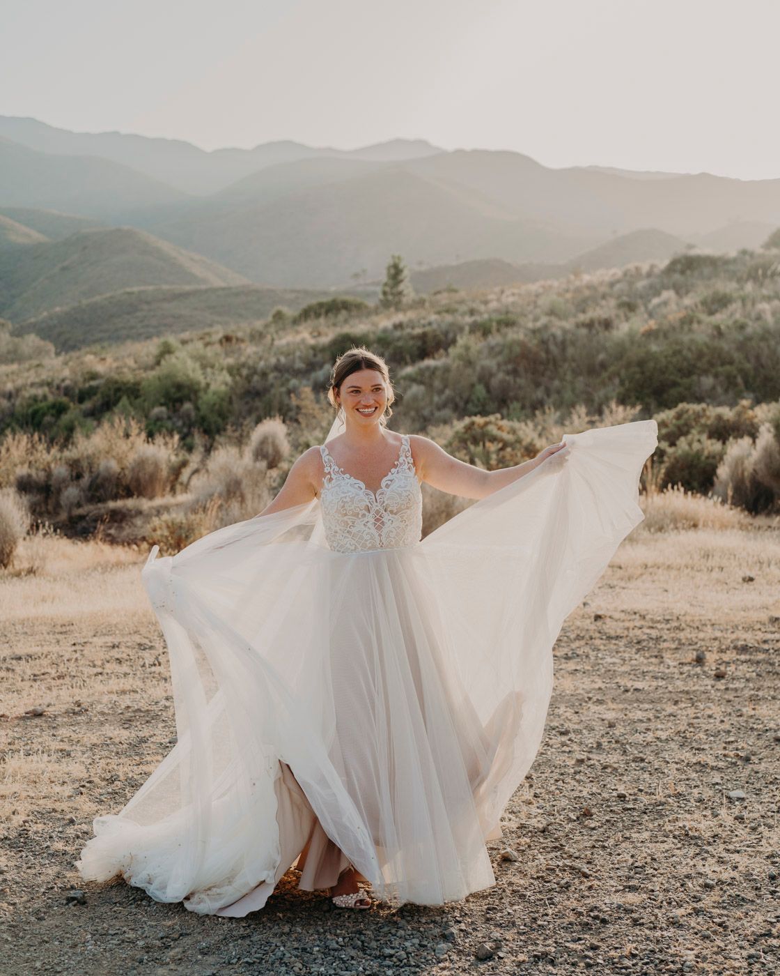 A flowing white wedding gown with fluttering fabric moves elegantly against a mountainous desert landscape at sunset.