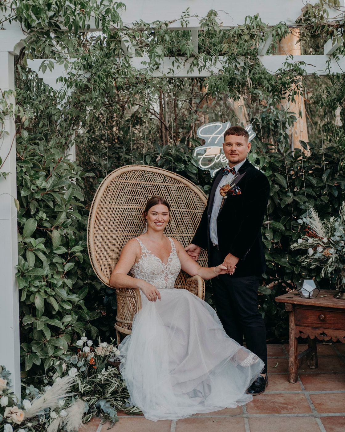 A wedding couple poses under a vine-covered pergola with a peacock chair and rustic decor.