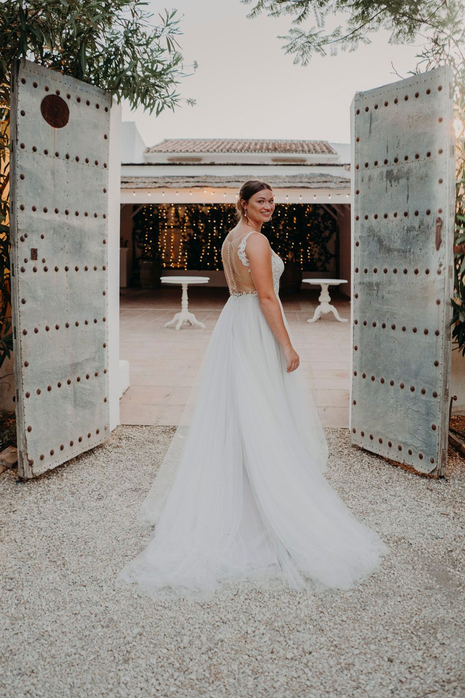 White wedding gown with flowing tulle skirt shown between rustic blue doors with twinkle lights in courtyard background.