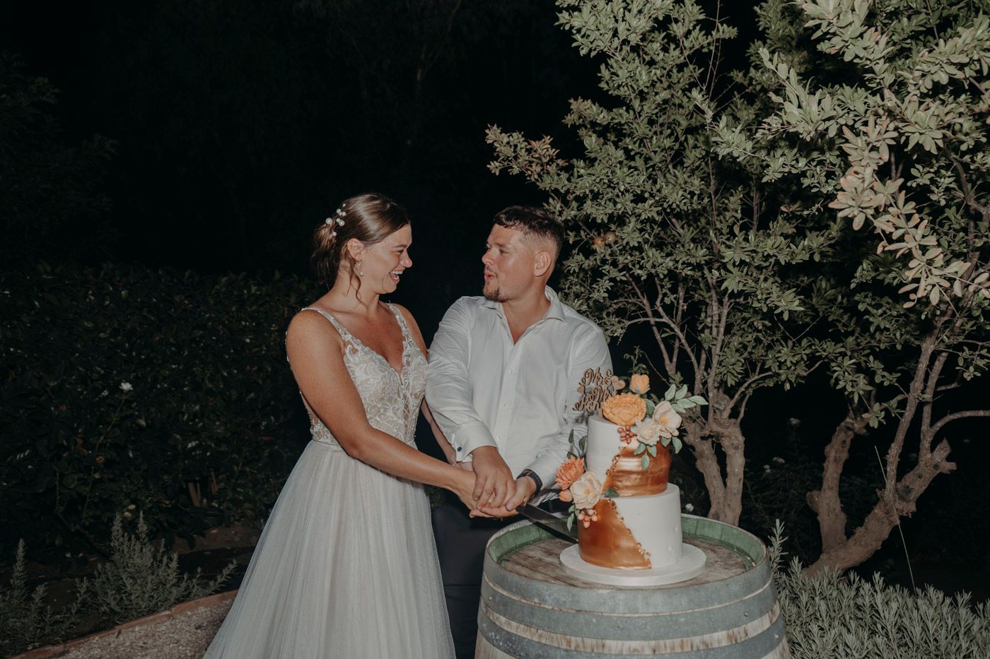 Couple shares a joyous moment near their rustic wedding cake displayed on a wine barrel at nighttime outdoor reception.