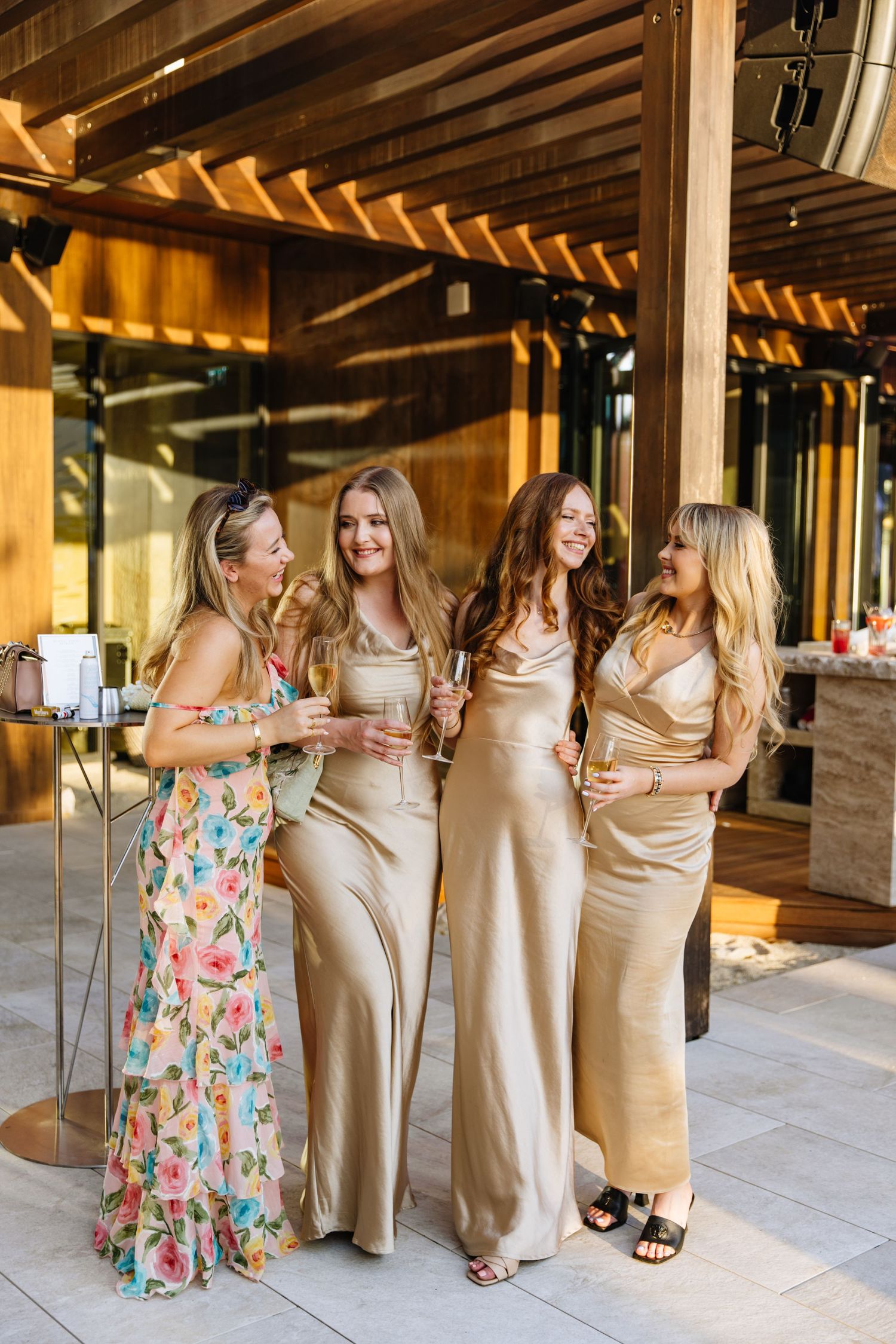A group of friends in formal attire laugh together at an elegant outdoor venue during a celebration.
