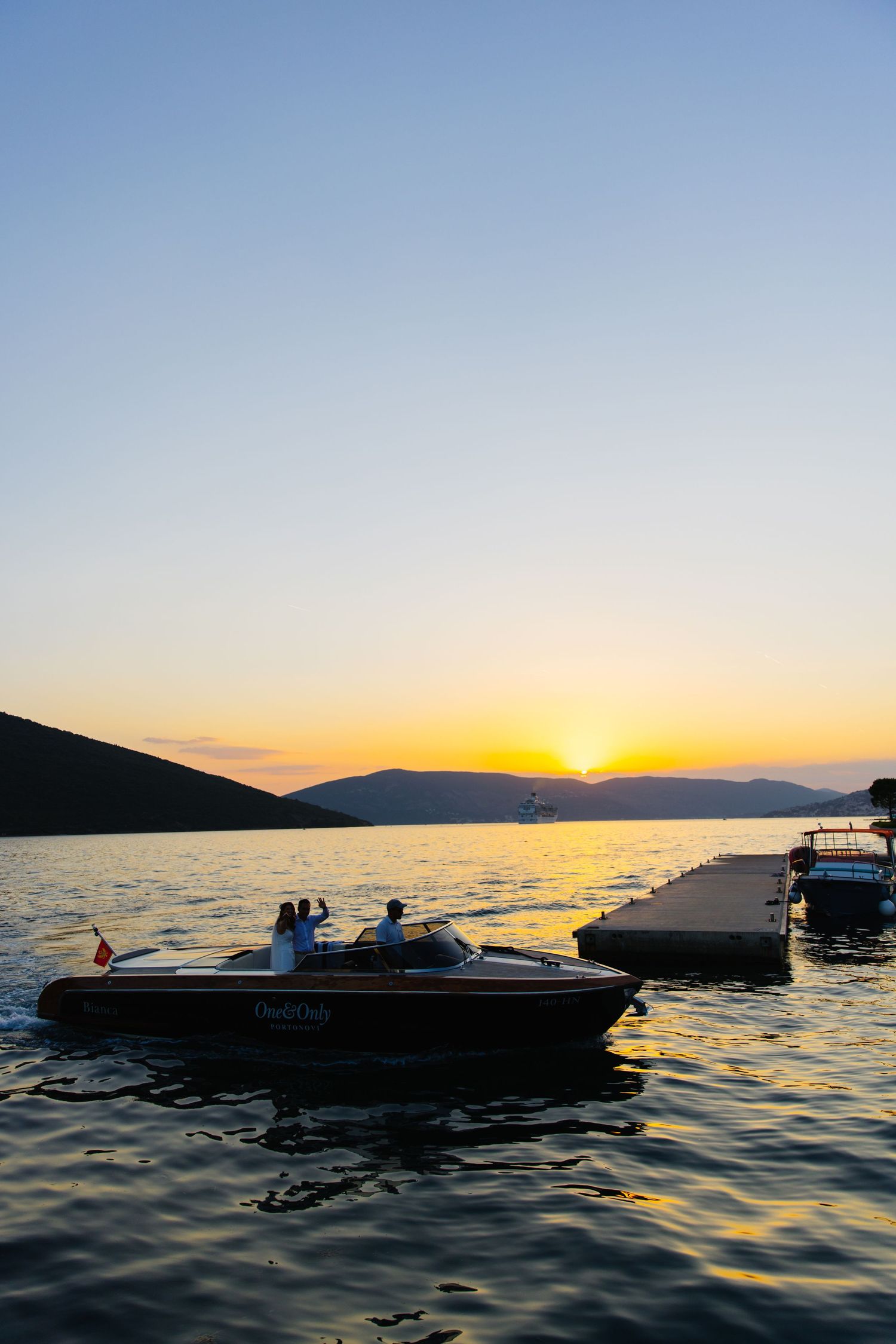 Silhouetted boat with passengers enjoying sunset over mountain lake with reflective waters and docked boats nearby.