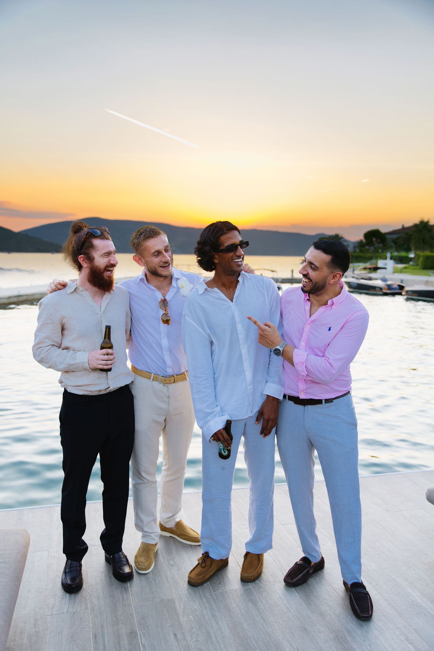 Four friends enjoying drinks and laughter on a dock during a beautiful sunset over the water.