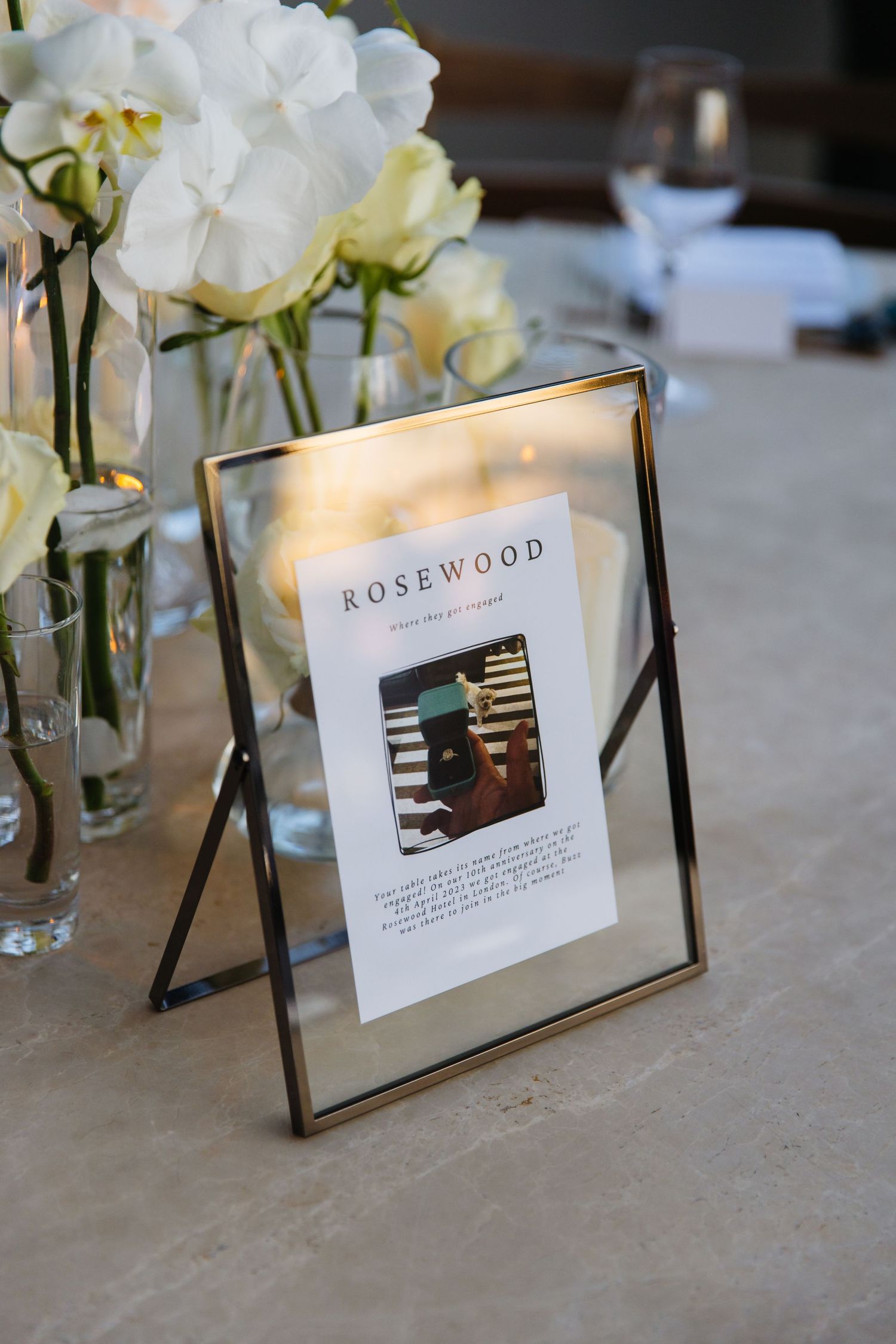 A Rosewood menu card in a glass frame sits on a table with delicate white and yellow floral centerpieces.
