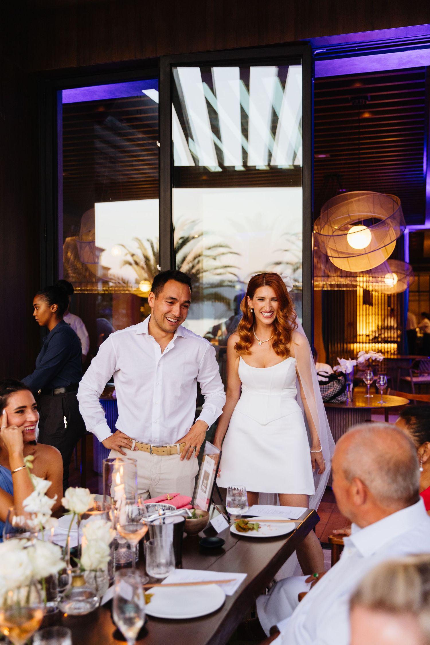 Wedding guests stand at an elegant dinner table in a modern restaurant with purple ambient lighting and decorative details.