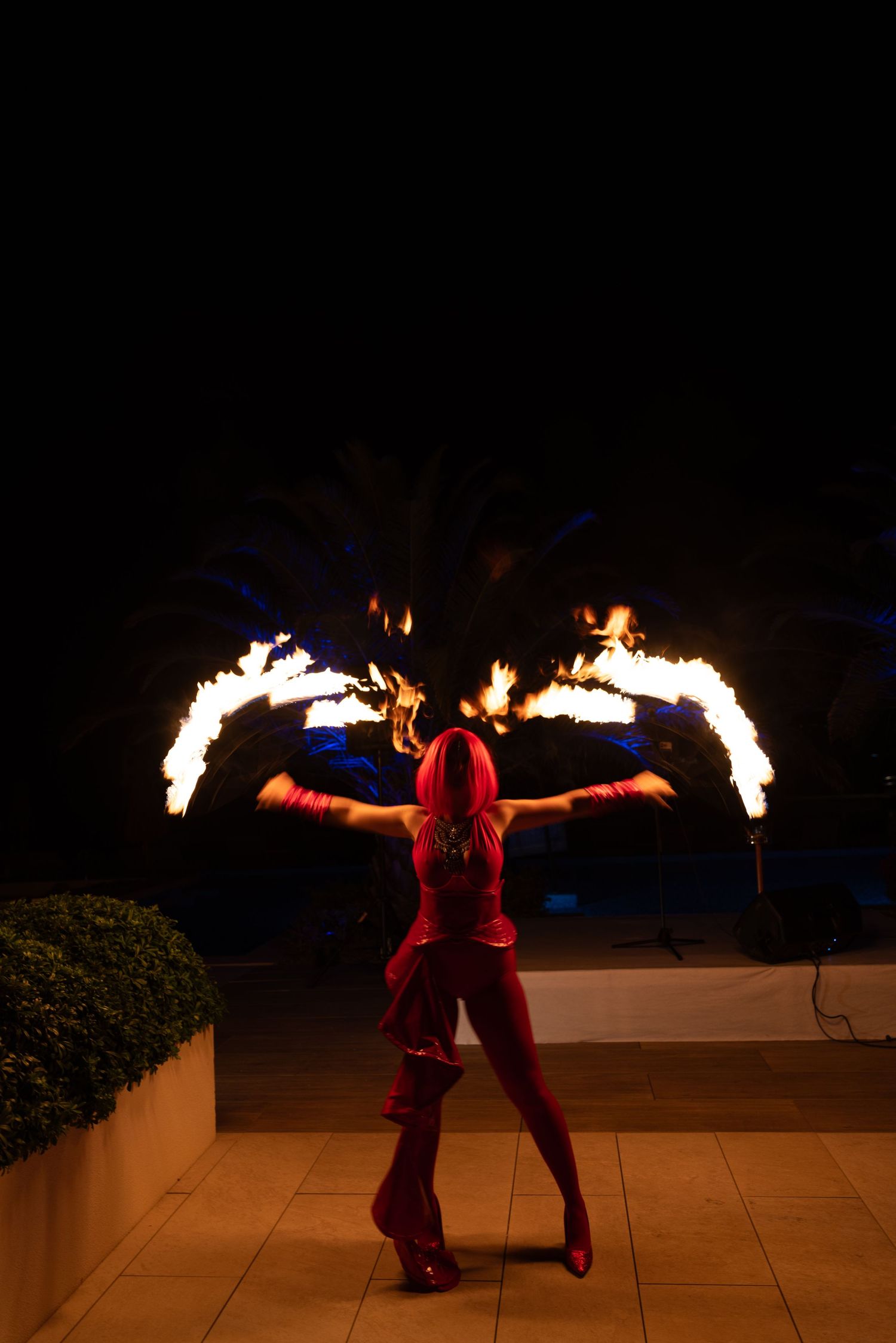 Fire performer in red costume spinning flaming batons at night during outdoor entertainment show.
