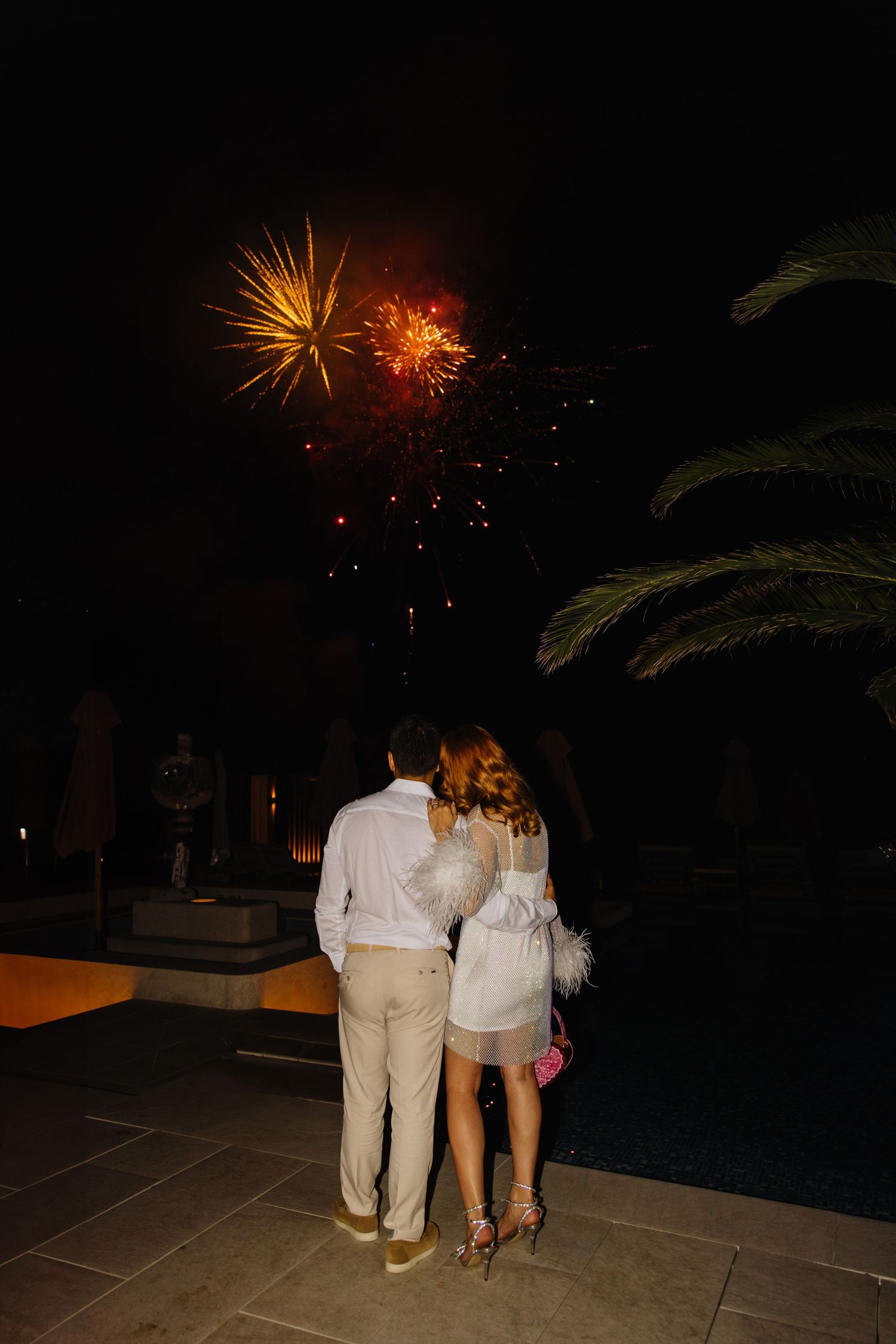 Couple watching fireworks display against dark sky with palm trees silhouetted in the foreground.