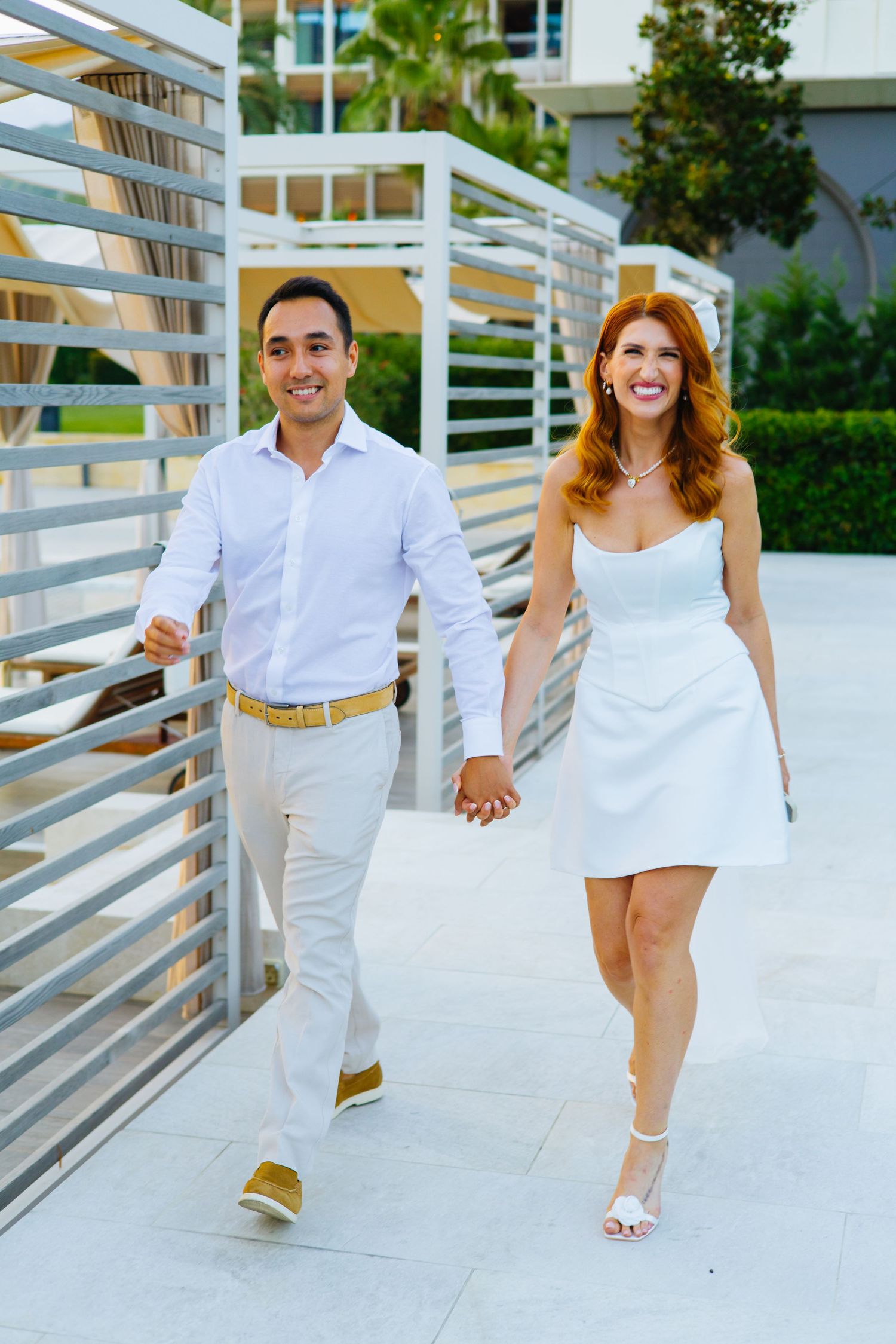 A couple in matching white outfits walks hand in hand on a modern walkway with white railings on a sunny day.