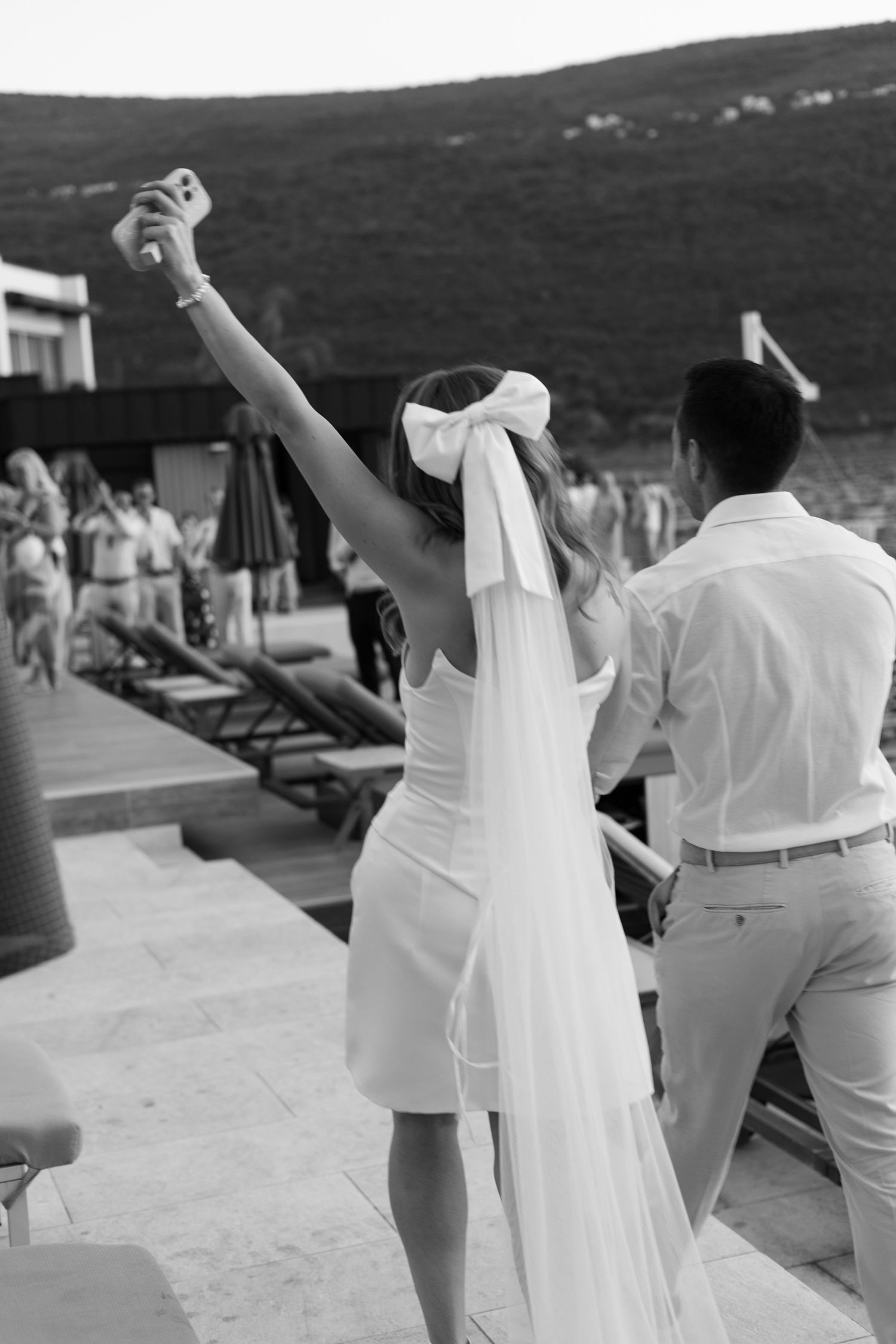 A bride in a short white dress and long veil celebrates with raised arms at an outdoor wedding venue.