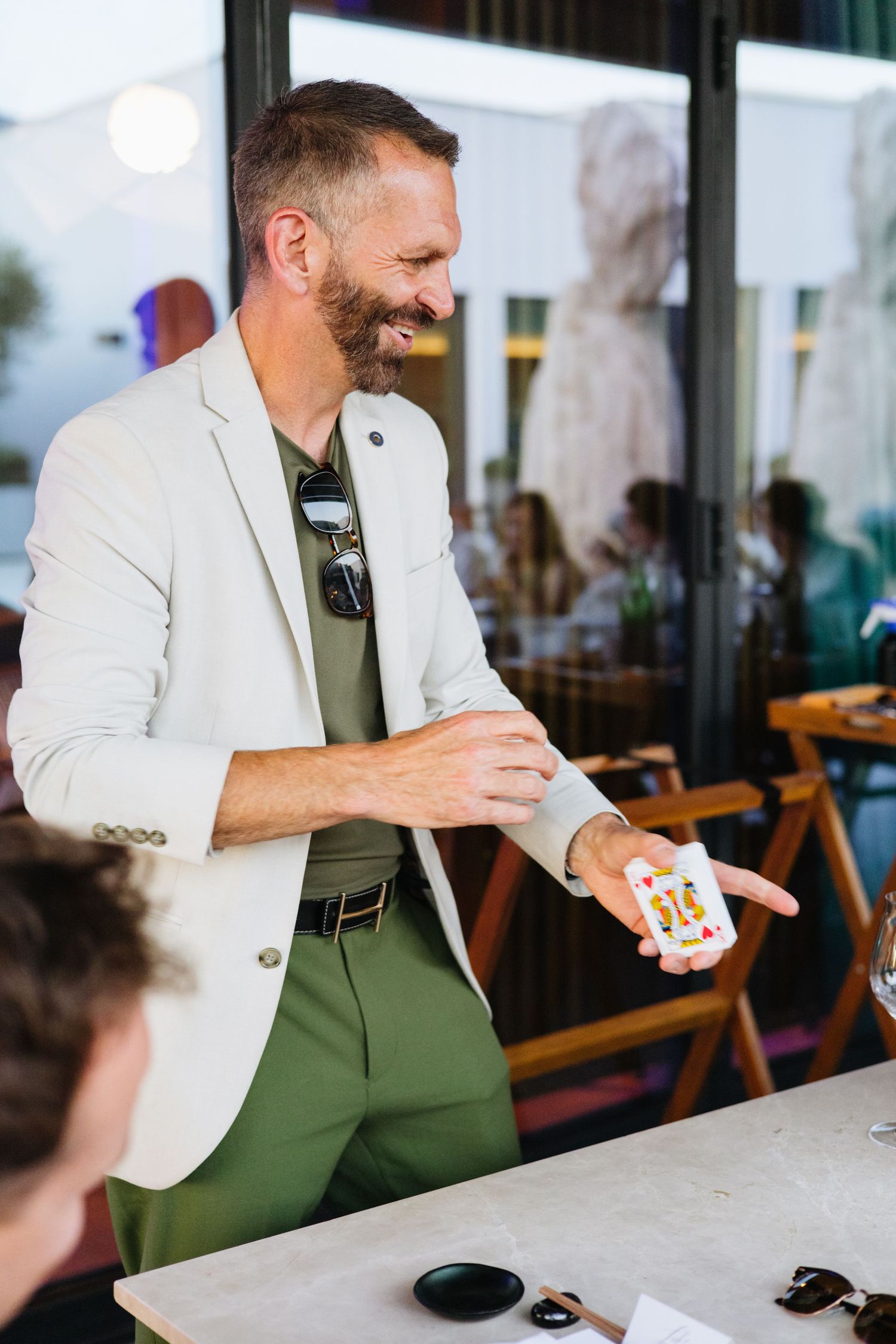 Professional in white blazer and green pants performing card trick at casual gathering in cafe setting.