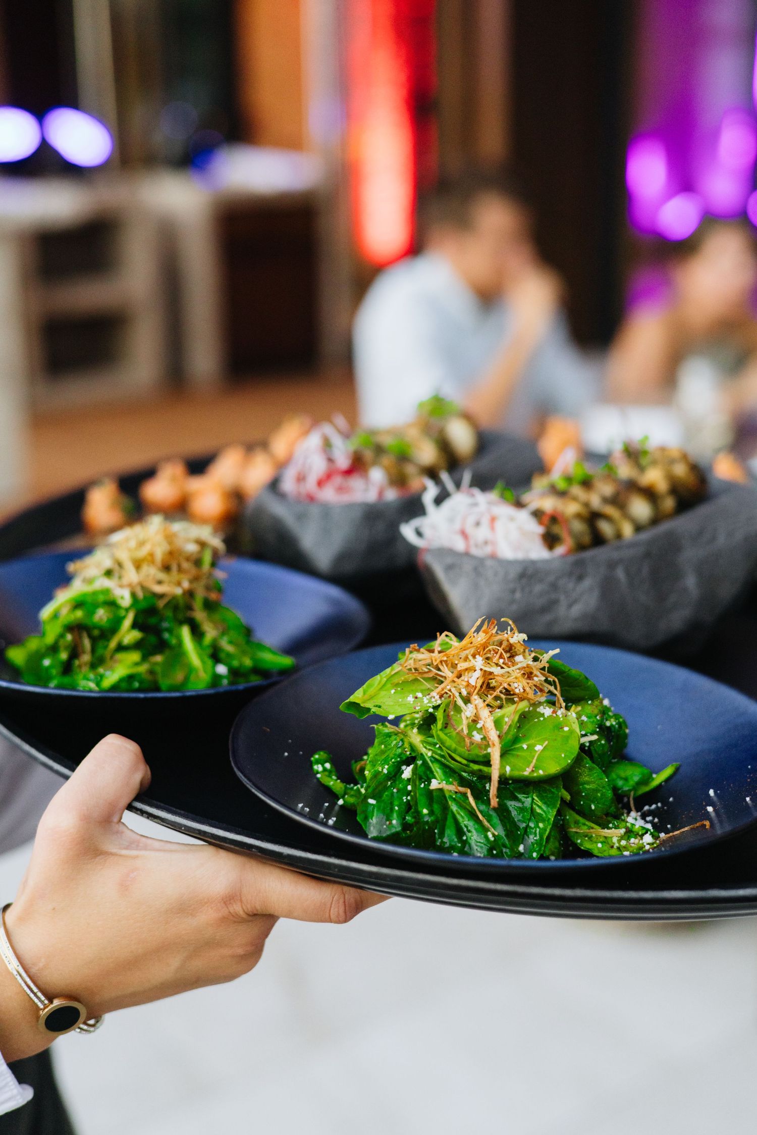 Server holding a tray of beautifully plated salads and dishes with colorful garnishes in a restaurant setting.