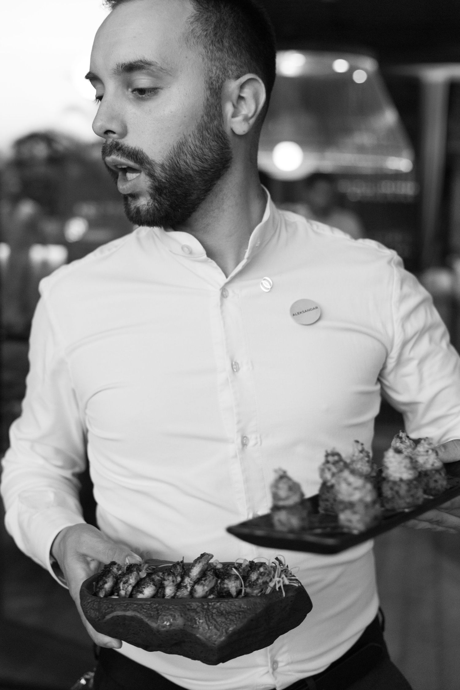 A server in a white button-down shirt holds stylish food dishes in a dimly lit restaurant setting.