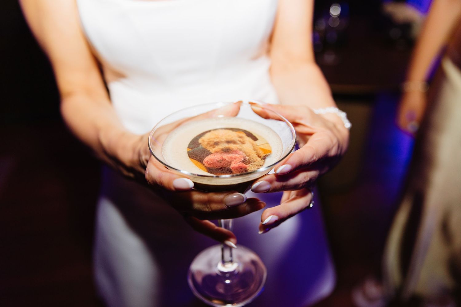 Hands holding an elegant martini glass with a decorative cocktail garnish against a purple-lit background.