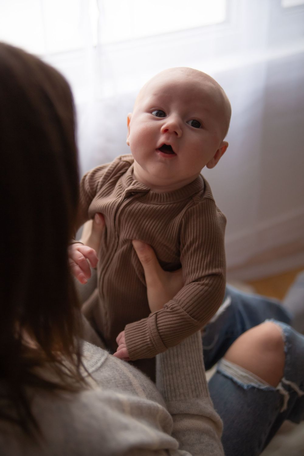 Baby wearing brown ribbed sweater is held by someone in ripped jeans against bright white background.