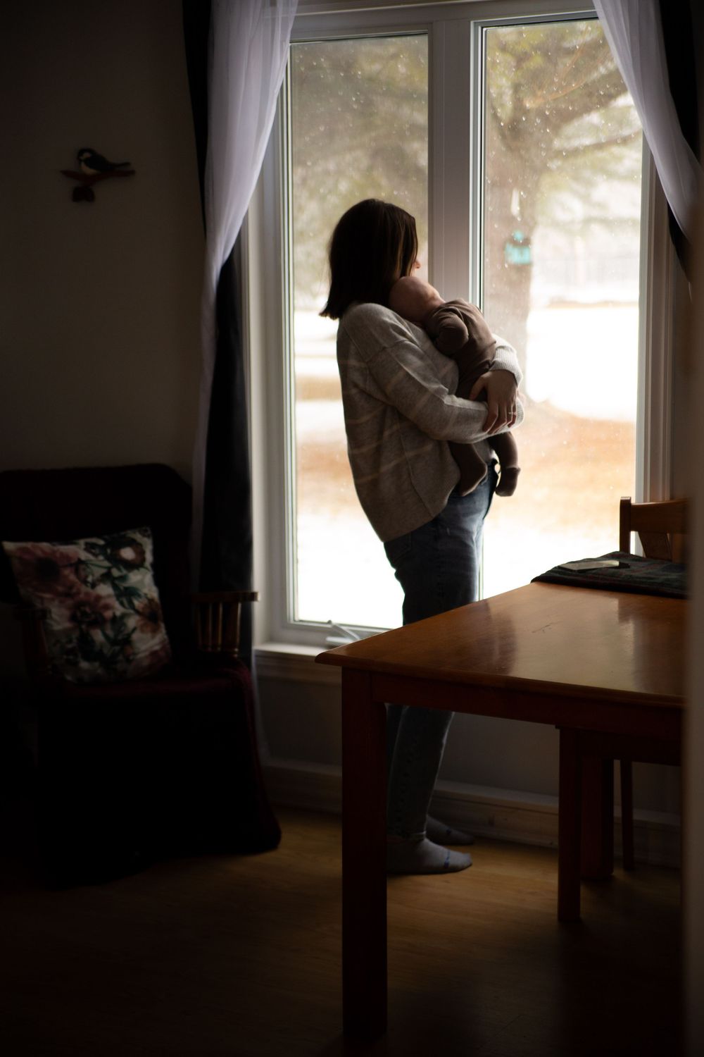 Silhouette of a figure holding a baby while standing by a window in a dimly lit living room.