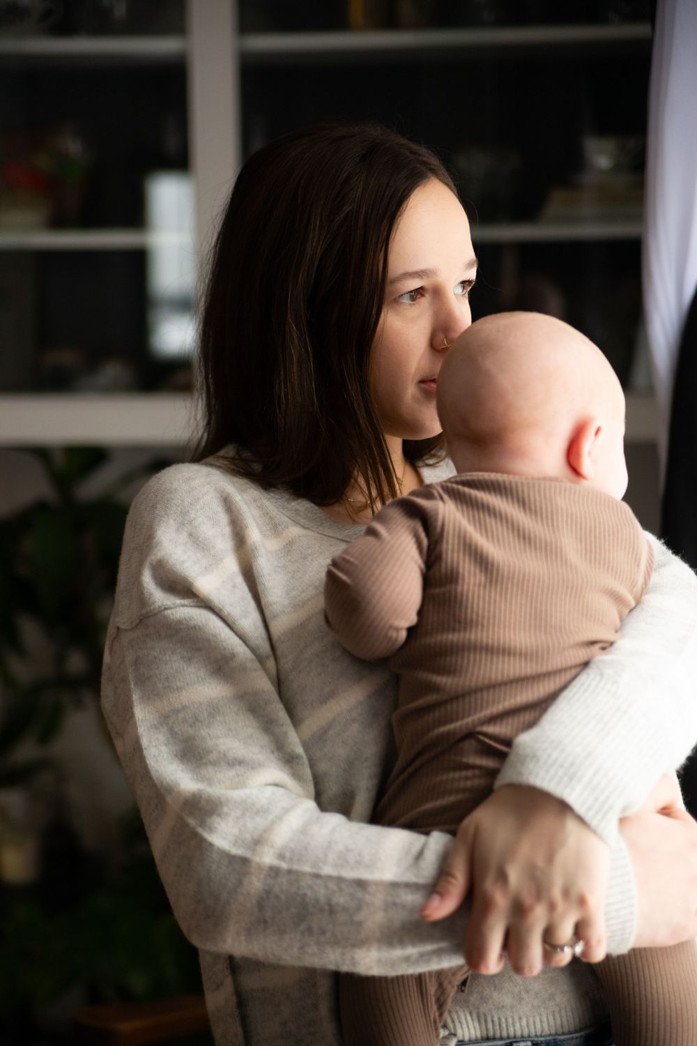 Tender moment of parent in cream sweater embracing infant in beige outfit by window.