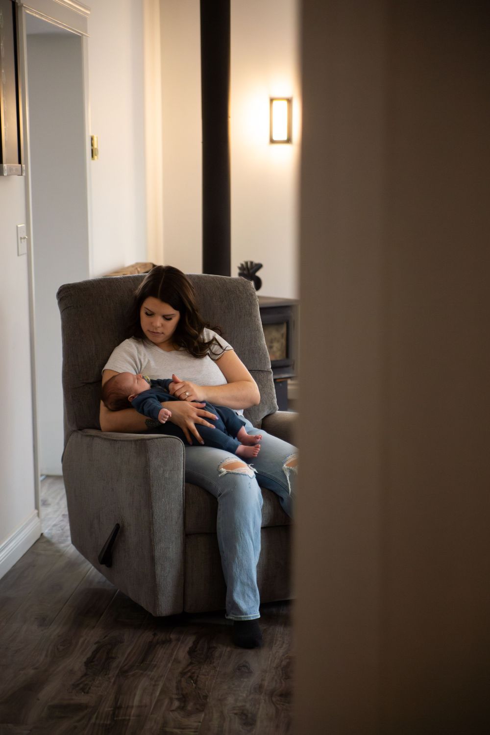 Someone sits in a grey recliner chair holding a small baby in a dimly lit room with hardwood floors.